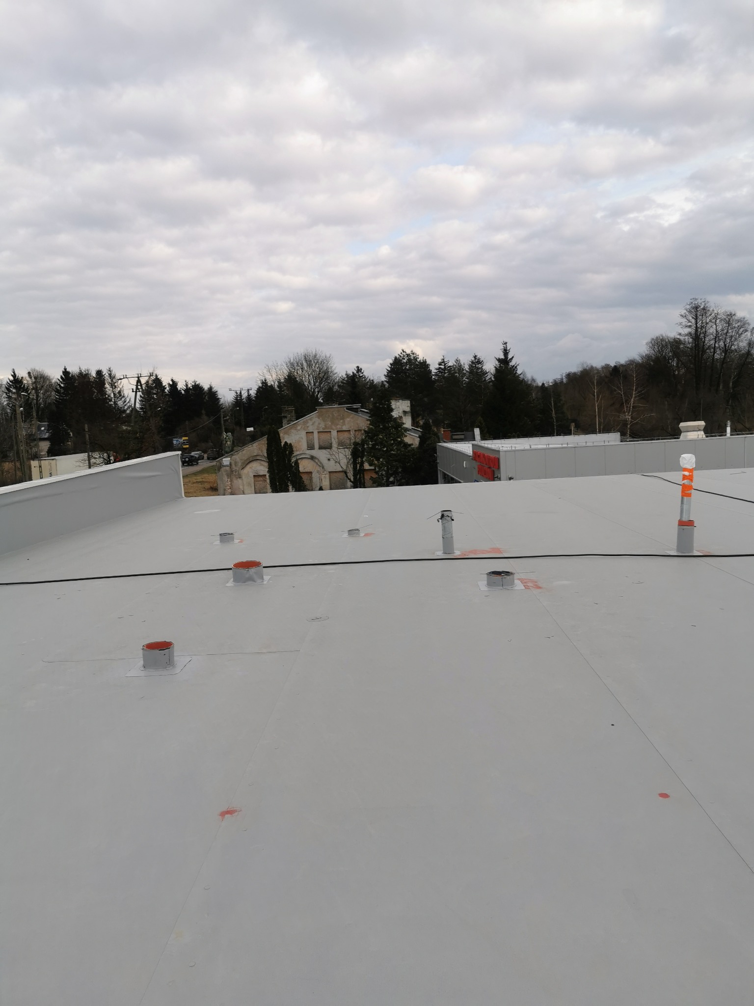 Płaski, szary dach przemysłowy z widocznymi kominami wentylacyjnymi i elementami instalacji, na tle zabudowań i drzew w pochmurny dzień.