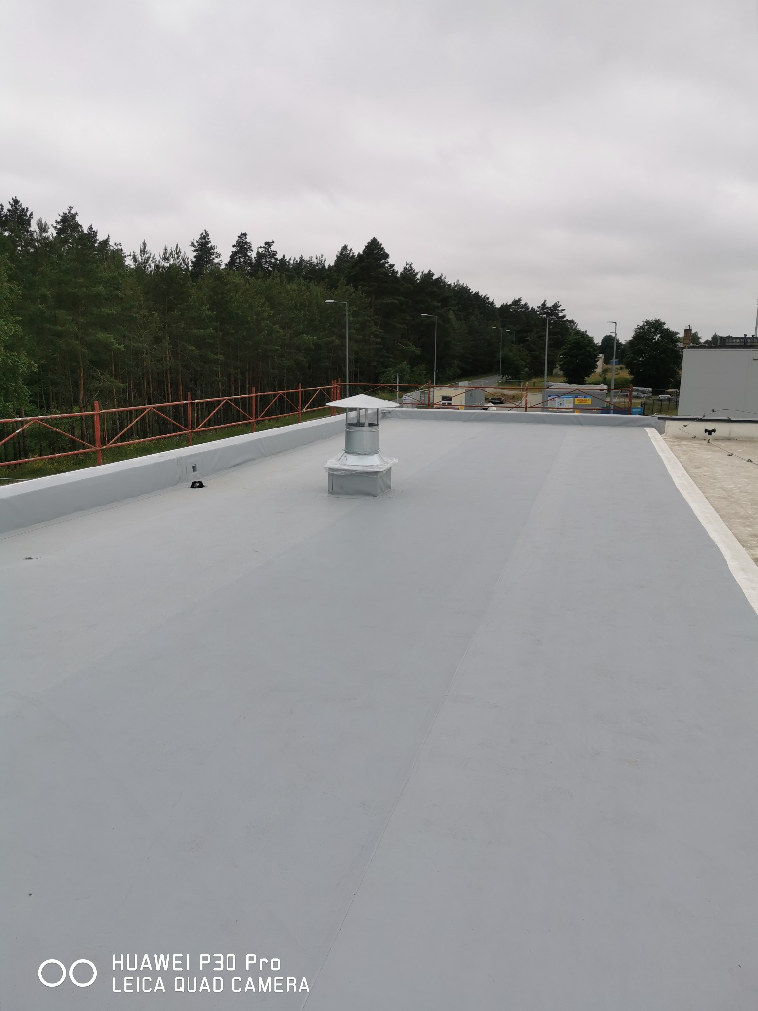 Szary, płaski dach pokryty papą termozgrzewalną z kominkiem wentylacyjnym pośrodku, widok z góry na tle lasu i pochmurnego nieba.