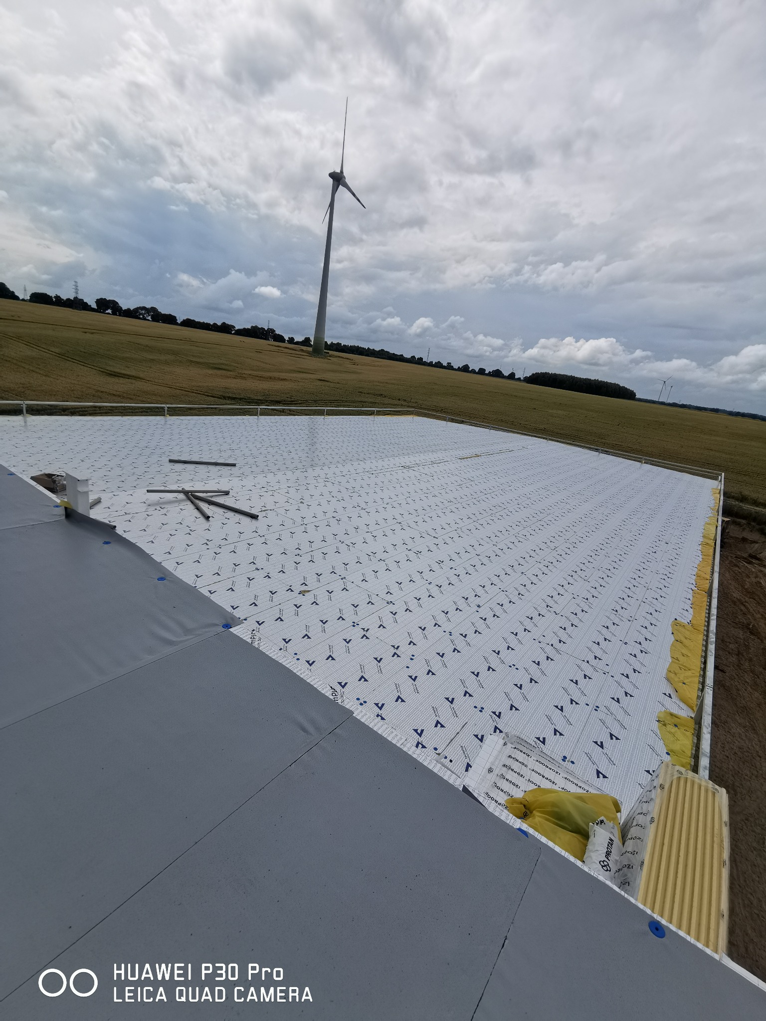 Widok z góry na dach pokryty membraną dachową z widocznymi elementami izolacji i turbiną wiatrową w tle.