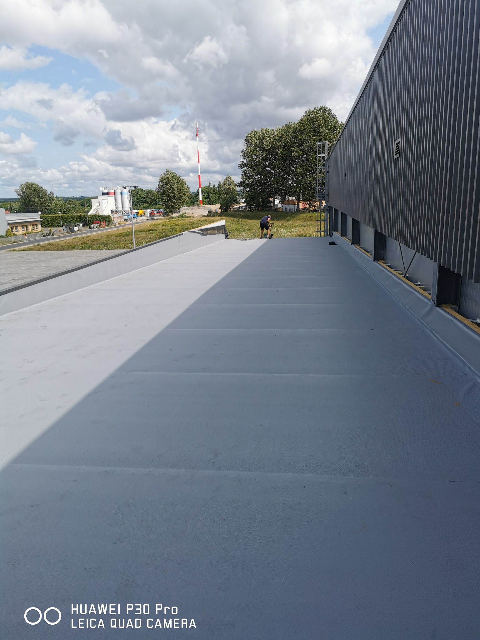 Szary, płaski dach przemysłowy z widocznym pracownikiem podczas inspekcji, metalowa ściana budynku w tle, częściowo zacieniony