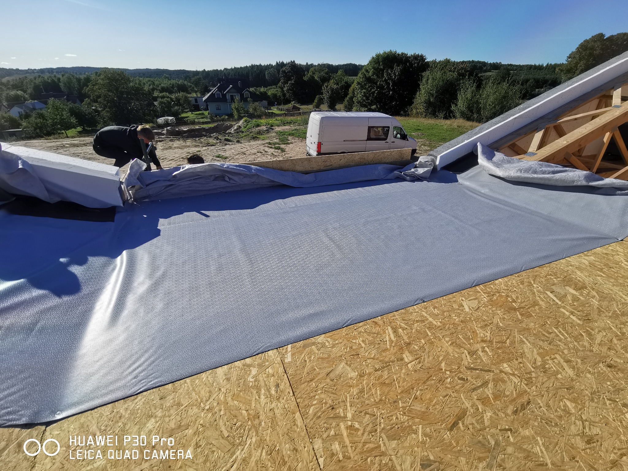 Prace dekarskie: Rozkładanie membrany dachowej na drewnianej konstrukcji dachu, widoczni dekarze i biały samochód dostawczy w tle.