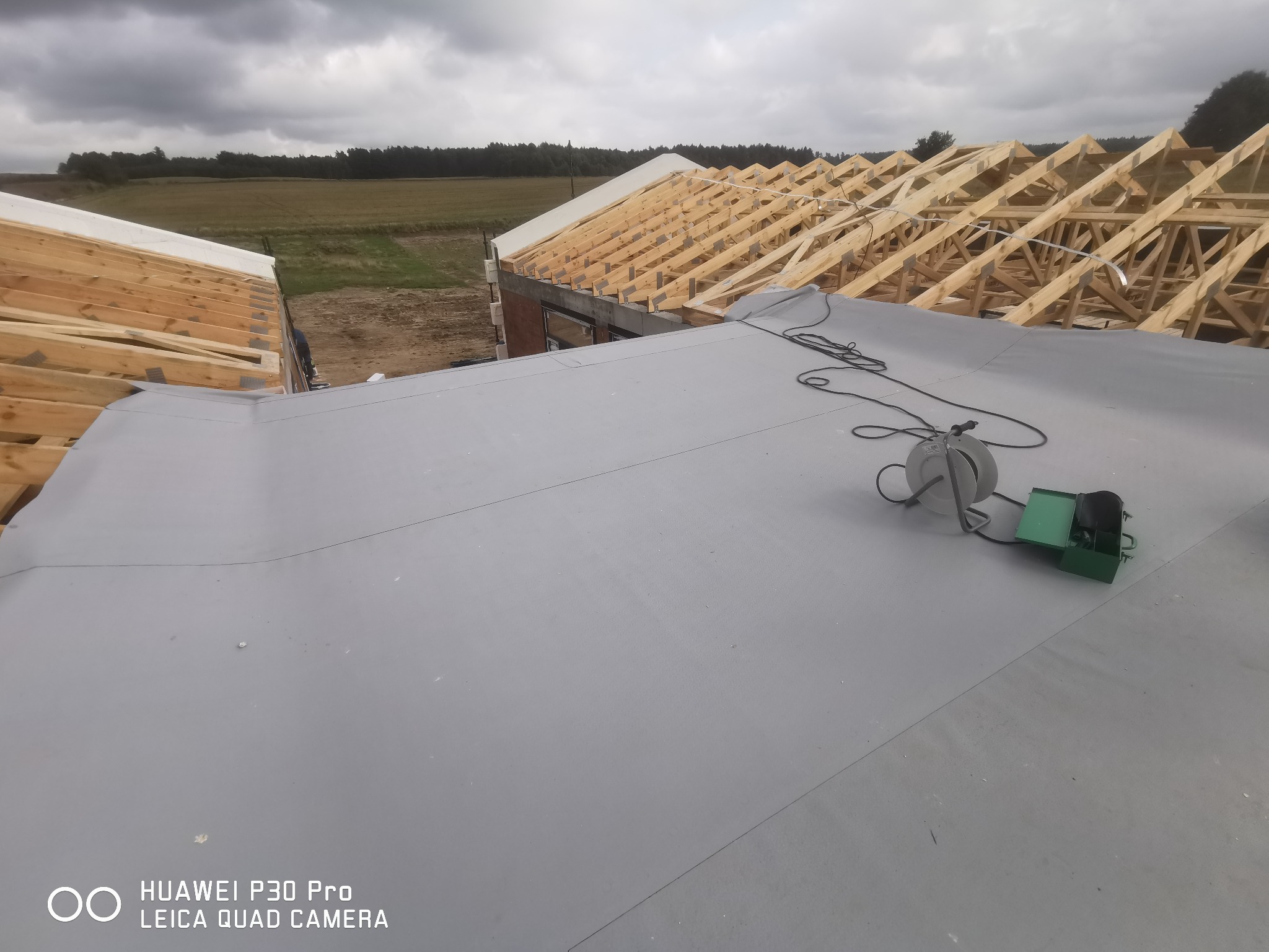 Szary dach z papy termozgrzewalnej w trakcie układania, widoczna konstrukcja drewniana dachu sąsiedniego budynku oraz narzędzia dekarskie: zgrzewarka i przedłużacz na dachu.