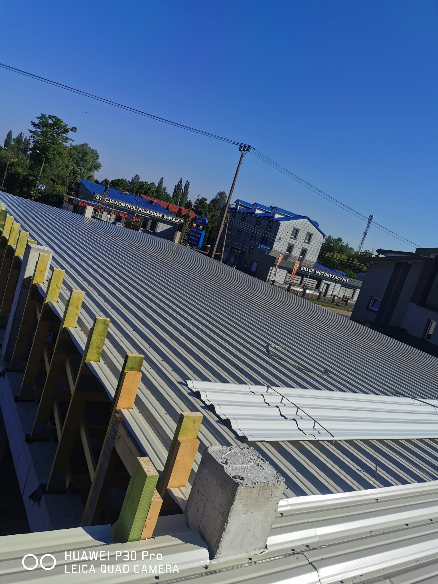 Montaż blachy trapezowej na dachu z drewnianą konstrukcją nośną, widok na budynki w tle w słoneczny dzień.