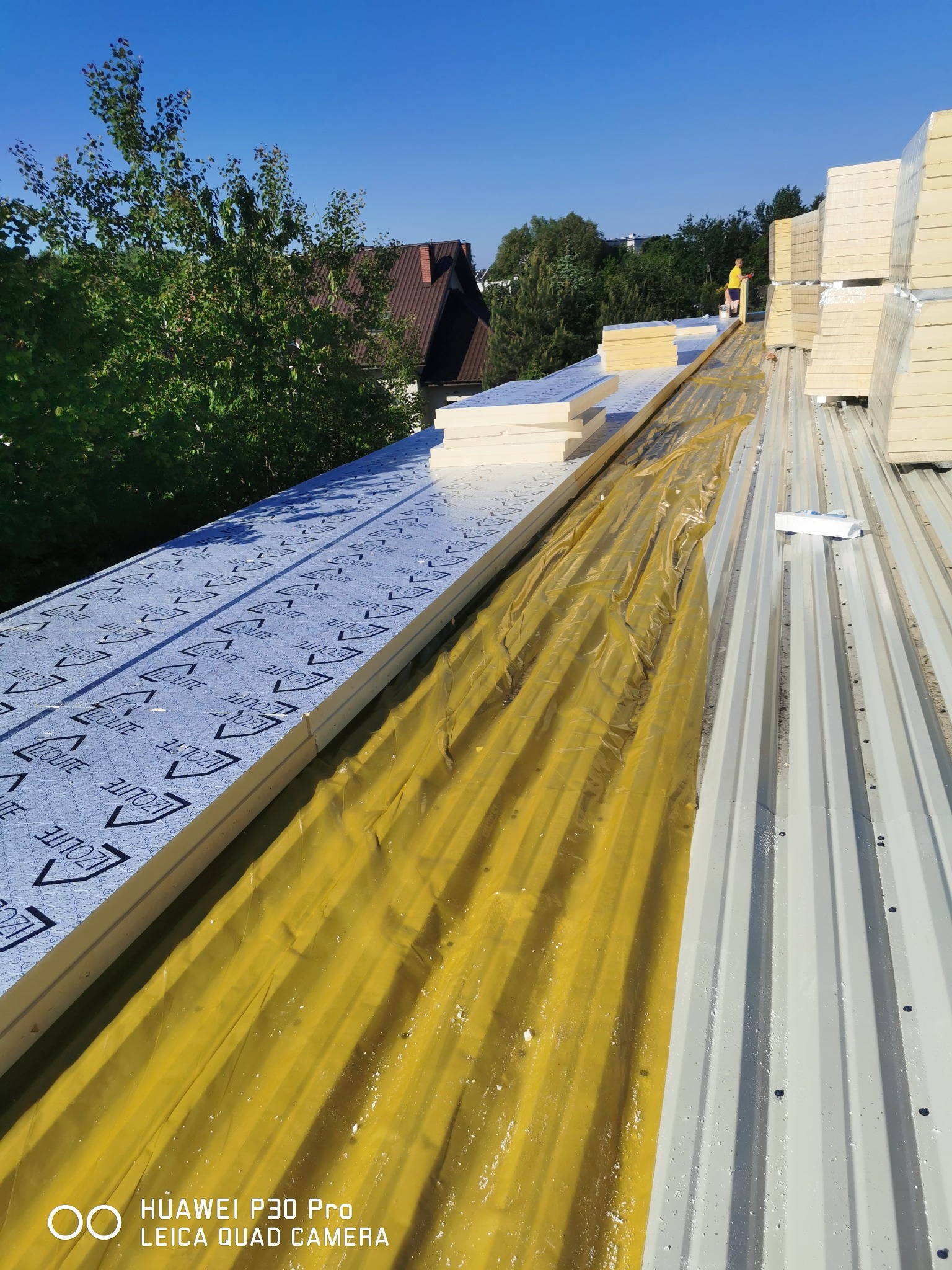 Montaż izolacji termicznej na dachu z blachy trapezowej, widoczna żółta folia paroizolacyjna i płyty izolacyjne z nadrukiem IZOlite, w tle pracownik w żółtej koszulce.
