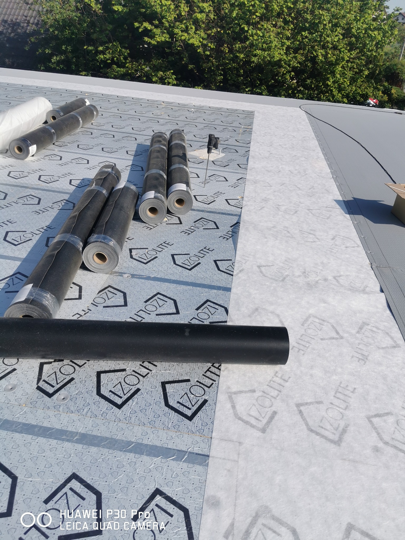 Rolki papy termozgrzewalnej Izolite na płaskim dachu, częściowo pokrytym białą membraną dachową, widoczne narzędzia dekarskie i drzewa w tle.