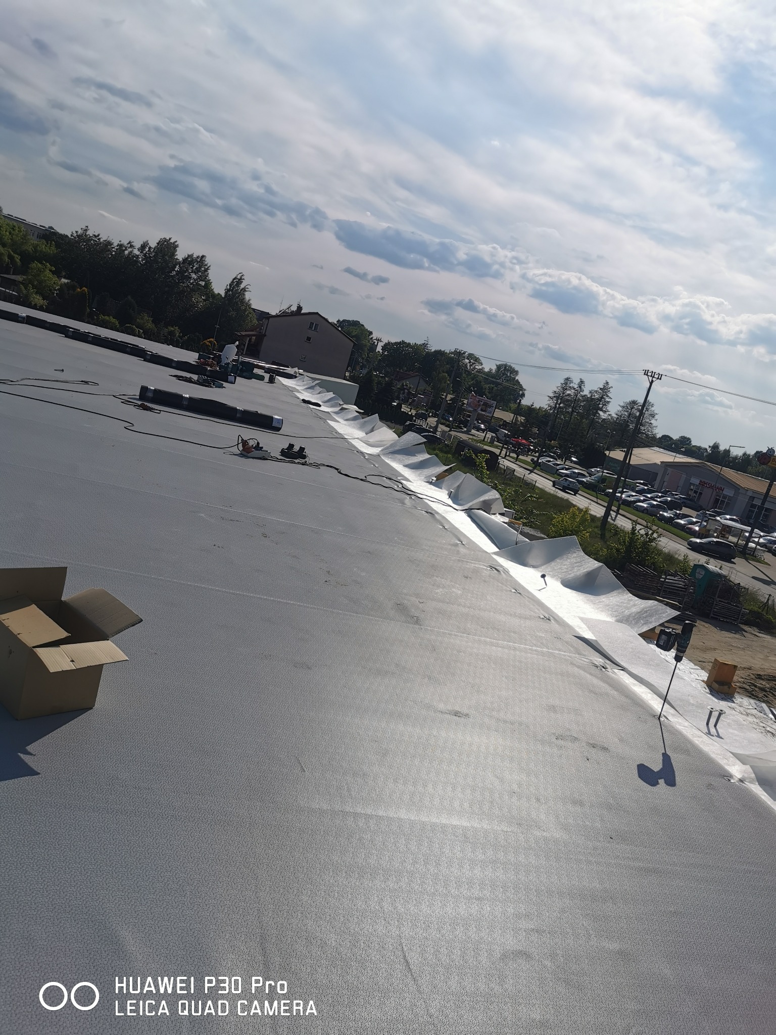 Szary płaski dach pokryty membraną, z otwartym kartonem w lewym dolnym rogu, na tle zabudowania i pochmurnego nieba.