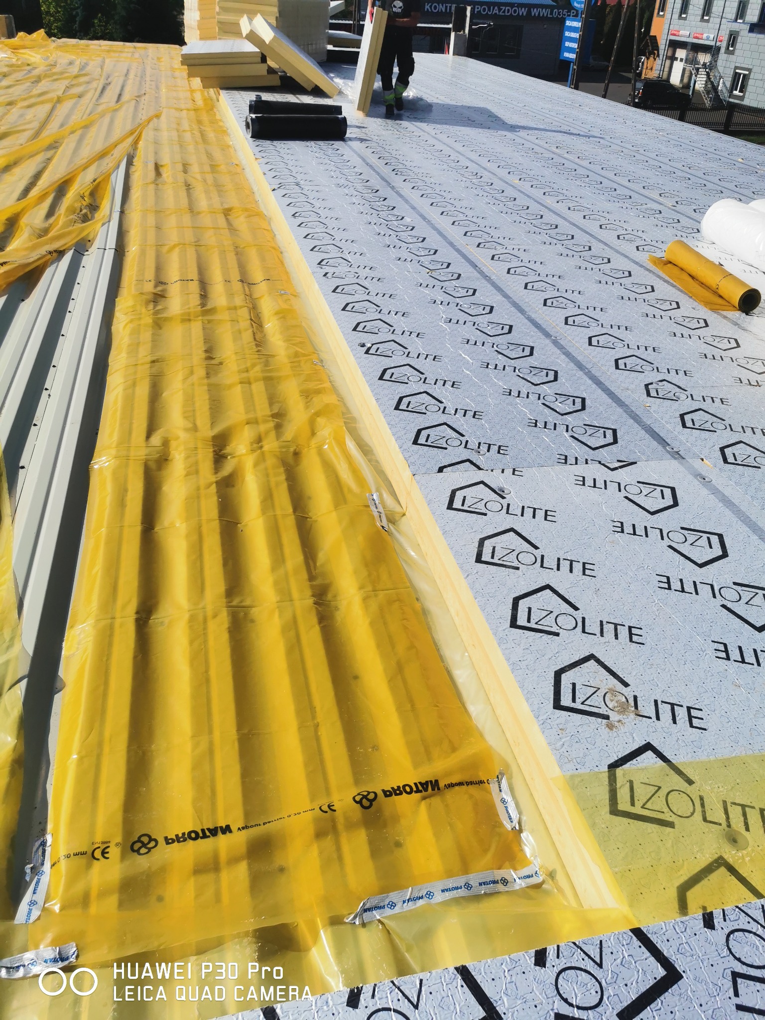 Układanie warstwy izolacyjnej Izolitte na blaszanym dachu, widoczne rolki folii ochronnej i płyty izolacyjne, częściowo zabezpieczone żółtą folią. Prace dekarskie w toku.