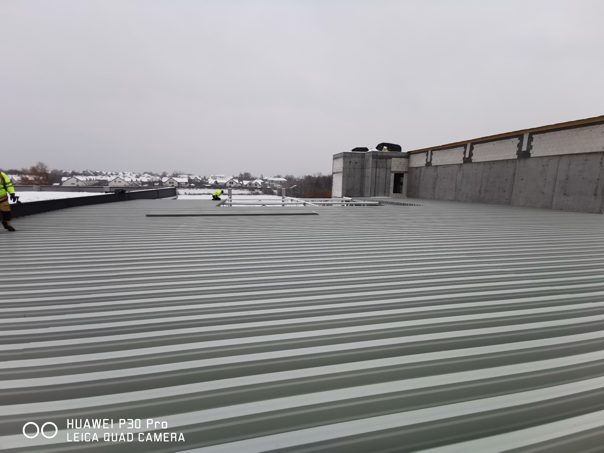 Montaż blachy trapezowej na dachu budynku przemysłowego, widoczni pracownicy w odblaskowych ubraniach, w tle zabudowa jednorodzinna.