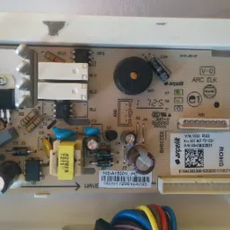 Płyta główna urządzenia AGD marki Arcelik, z widocznymi elementami elektronicznymi, takimi jak kondensatory, rezystory i układy scalone, umieszczona w białej obudowie.