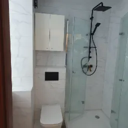 Nowoczesna łazienka z białymi płytkami imitującymi marmur, kabiną prysznicową z czarną armaturą, podwieszaną toaletą i szafką nad nią, oświetleniem punktowym.