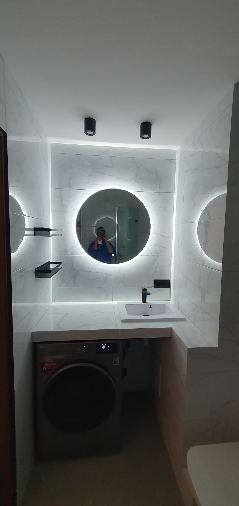 Nowoczesna łazienka w Brodnicy z marmurowymi płytkami, okrągłym lustrem z podświetleniem LED, białą umywalką z czarną baterią i pralką pod blatem.