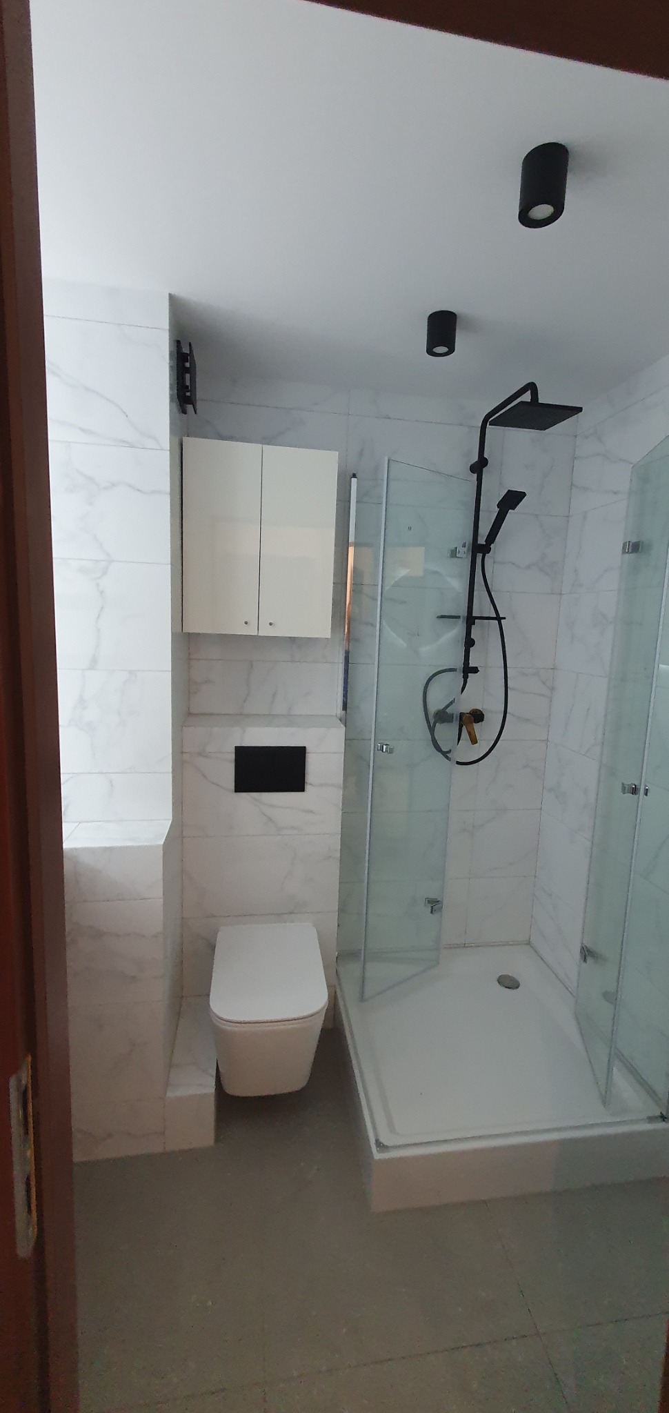 Nowoczesna łazienka z białymi płytkami imitującymi marmur, kabiną prysznicową z czarną armaturą, podwieszaną toaletą i szafką nad nią, oświetleniem punktowym.