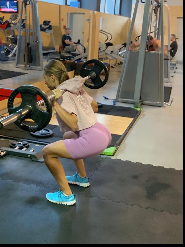 Kobieta w różowych spodenkach i niebieskich butach wykonuje przysiad ze sztangą na plecach w siłowni. W tle widać inne osoby ćwiczące na maszynach.