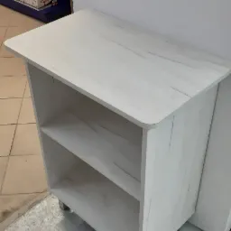 Biała szafka z półkami, imitująca strukturę drewna, z zaokrąglonymi krawędziami blatu i metalowymi nóżkami, ustawiona na szarej podłodze w pomieszczeniu z białymi ścianami.