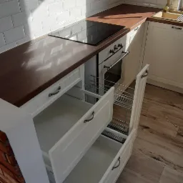 Jasna kuchnia z drewnianym blatem, widoczne otwarte szuflady z metalowymi koszami, białe kafelki na ścianie i drewniana podłoga.