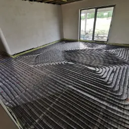 Instalacja ogrzewania podłogowego w surowym wnętrzu, widoczne rury grzewcze ułożone na izolacji z siatką, ściany w stanie deweloperskim, duże okno tarasowe z widokiem na zewnątrz.