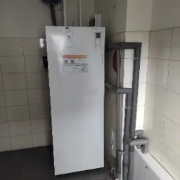 Biała pompa ciepła Atlantic zamontowana w pomieszczeniu z kafelkami, widoczne rury z izolacją termiczną.