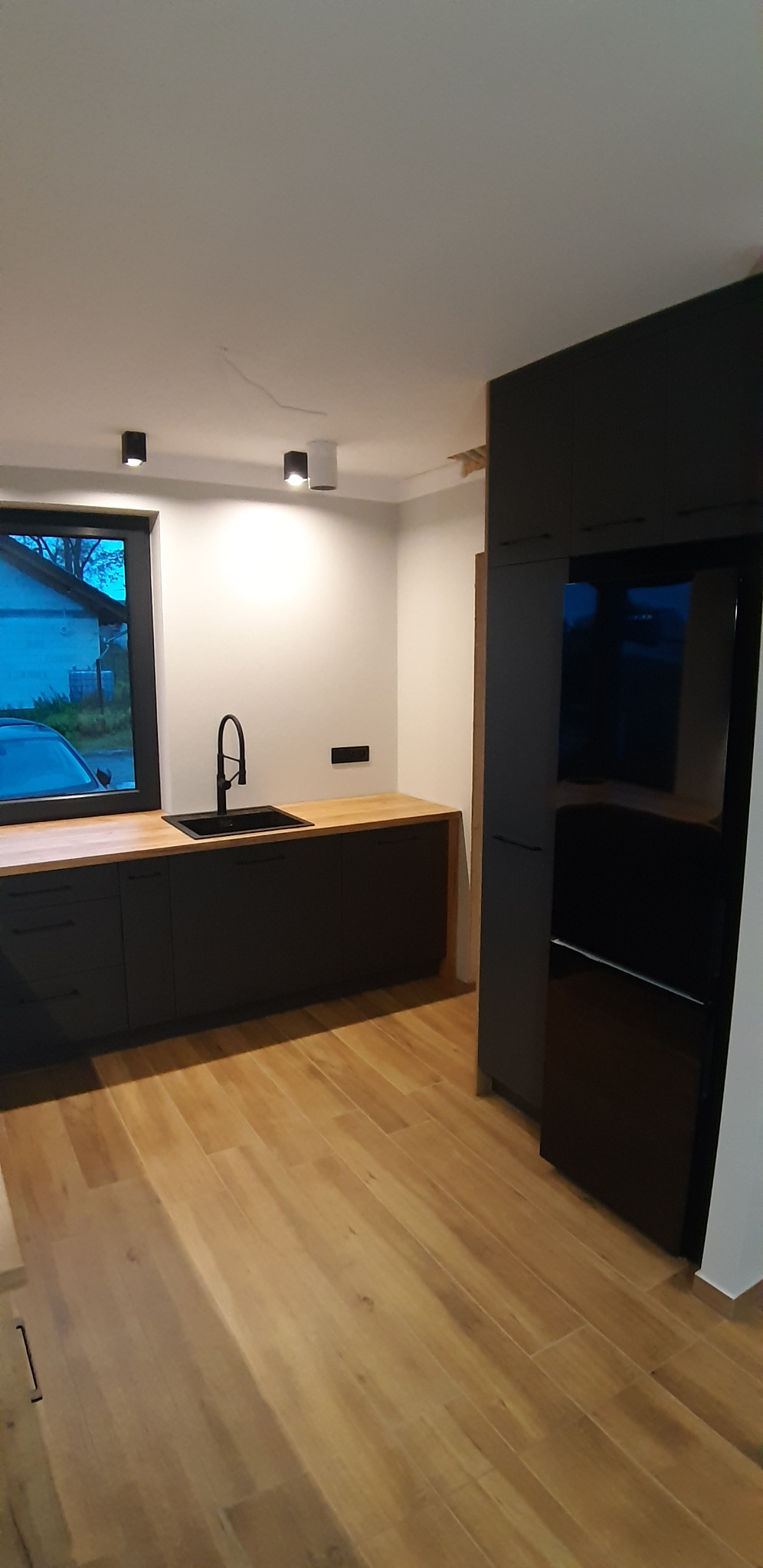 Nowoczesna kuchnia z czarnymi szafkami, drewnianym blatem i podłogą imitującą drewno, widok na okno zza zlewu.