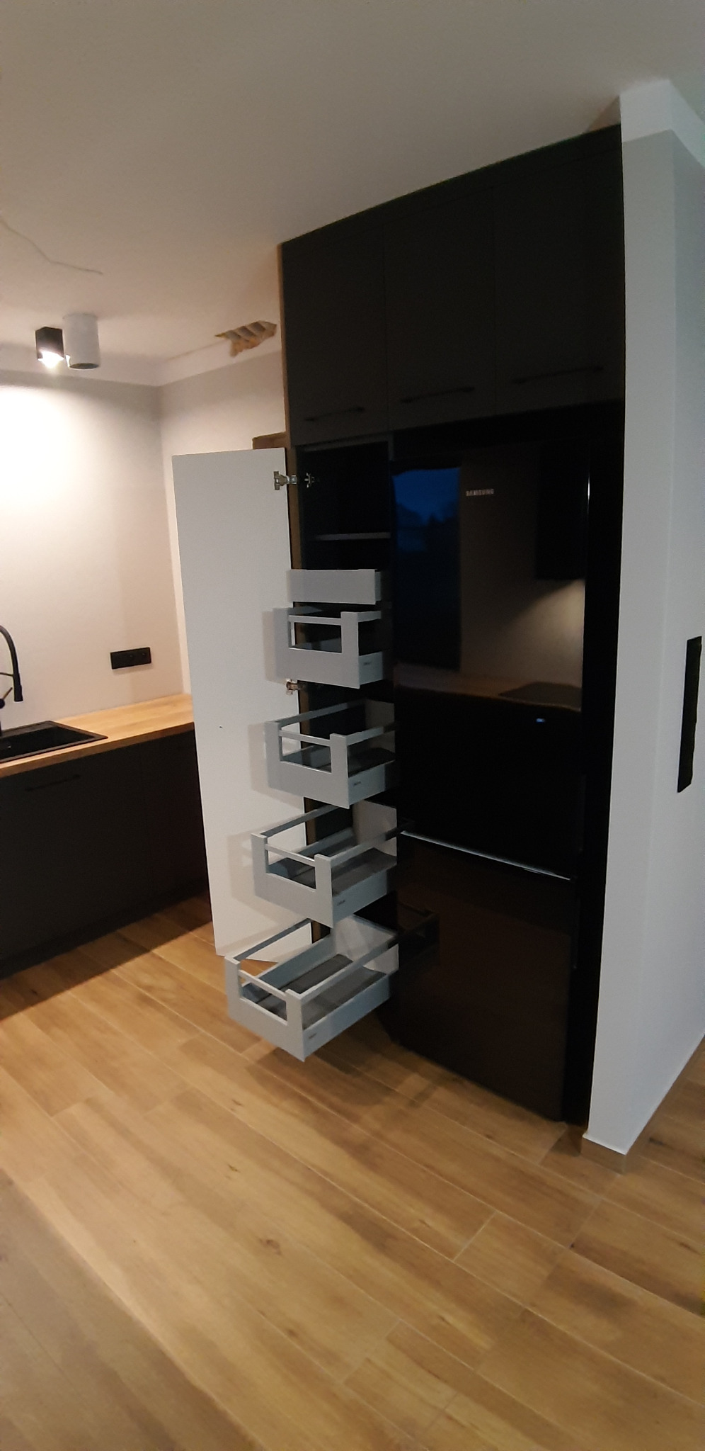 Ciemna kuchnia z otwartą szafką cargo z szufladami, widoczny fragment blatu i zlewu, drewniana podłoga.