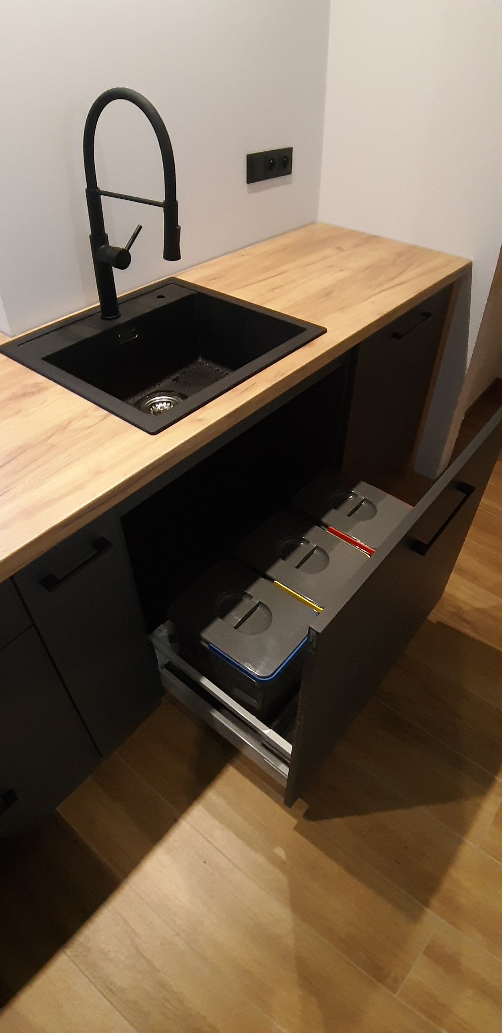 Nowoczesna kuchnia z blatem imitującym drewno, czarnym zlewem i baterią, szuflada z sortownikami na śmieci wysunięta.