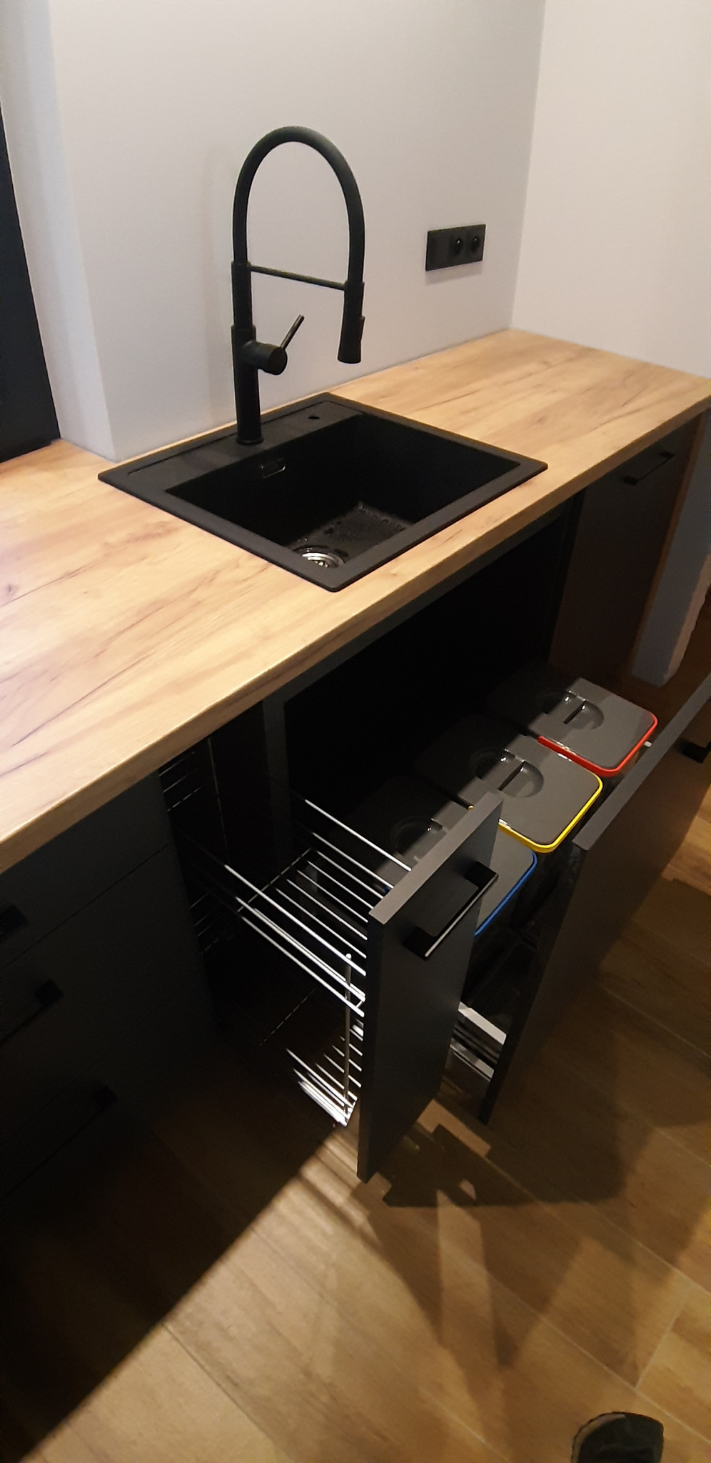 Nowoczesna kuchnia z czarnym zlewem i baterią, blat z wzorem drewna, wysuwany kosz na śmieci z kolorowymi pokrywami.