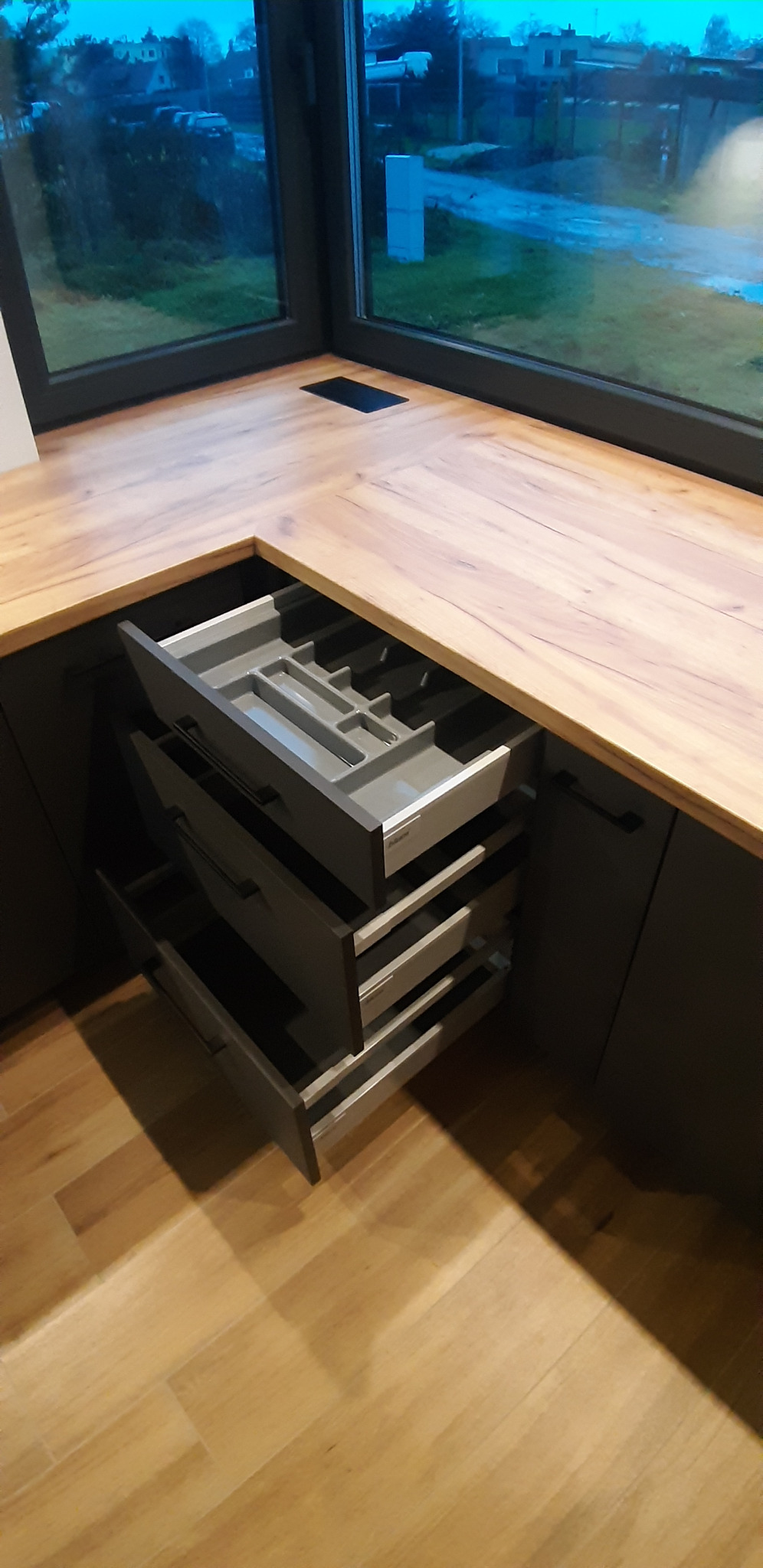 Szare, otwarte szuflady z przegródkami na sztućce w nowoczesnej kuchni z drewnianym blatem roboczym i oknem z widokiem na zewnątrz.