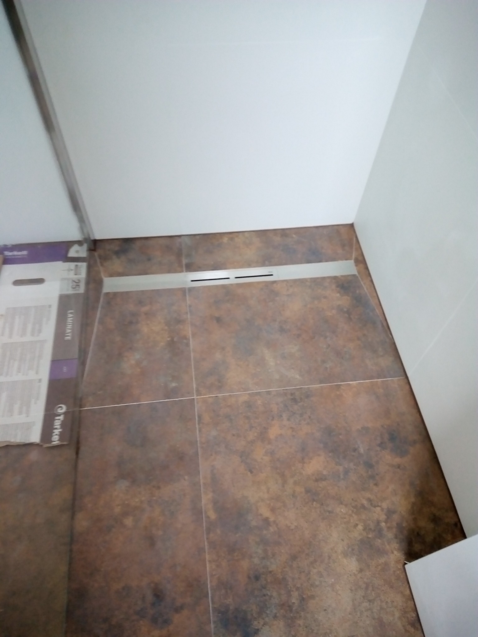 Ułożona brązowa terakota w kabinie prysznicowej z odpływem liniowym, widoczny fragment opakowania płytek Tarkett Laminate.