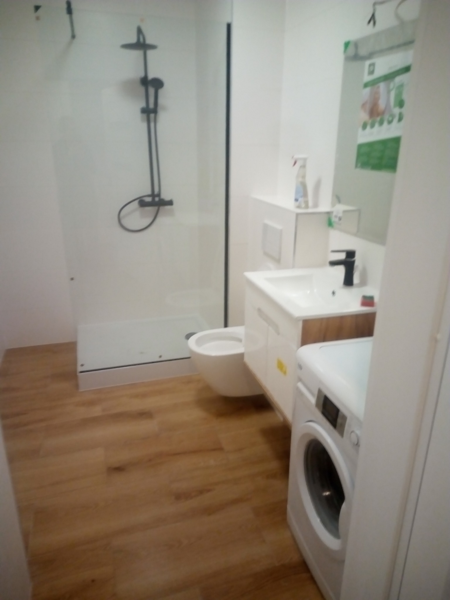 Nowoczesna łazienka z kabiną prysznicową typu walk-in z czarnym zestawem natryskowym, umywalką z szafką z elementami drewnopodobnymi, toaletą i pralką.