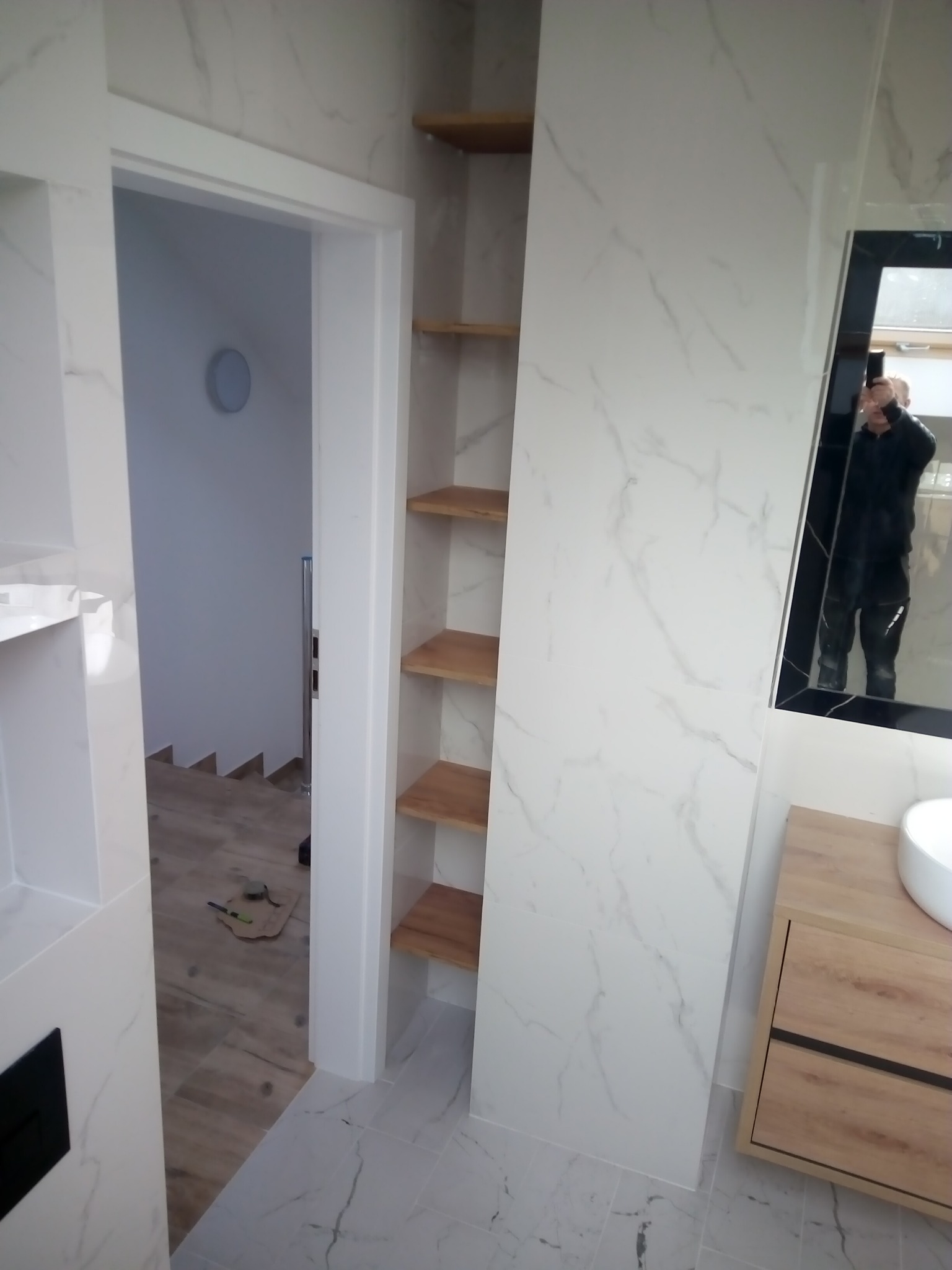 Wykończona łazienka z płytkami imitującymi marmur, drewnianymi półkami w zabudowie i szafką pod umywalkę w odcieniach drewna, widoczne narzędzia na podłodze w przejściu do innego pomieszczenia.