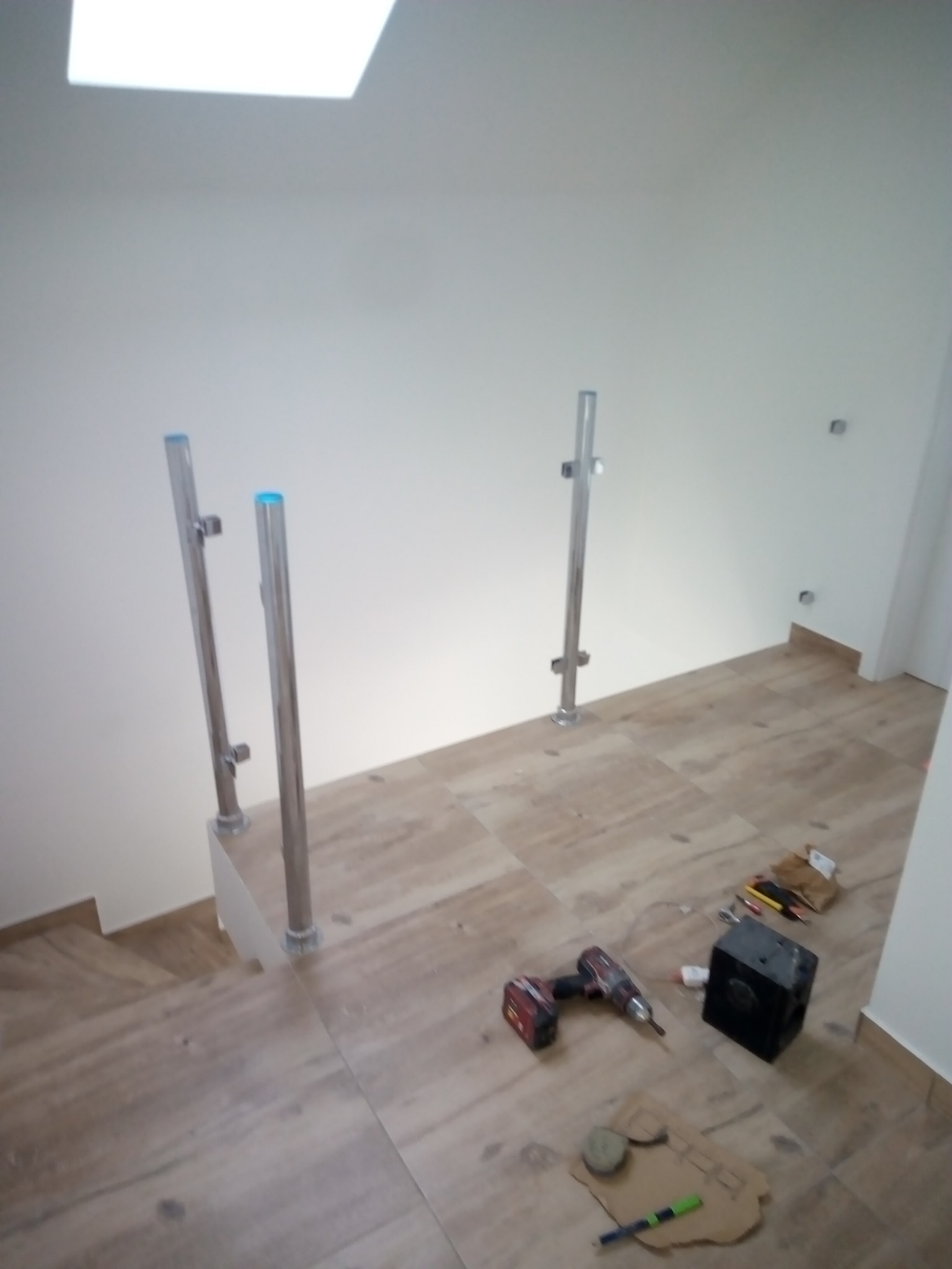 Montaż balustrady schodowej z metalowymi słupkami na schodach wyłożonych płytkami imitującymi drewno. Widoczne narzędzia pracy: wiertarka, poziomica, kartonowy szablon.