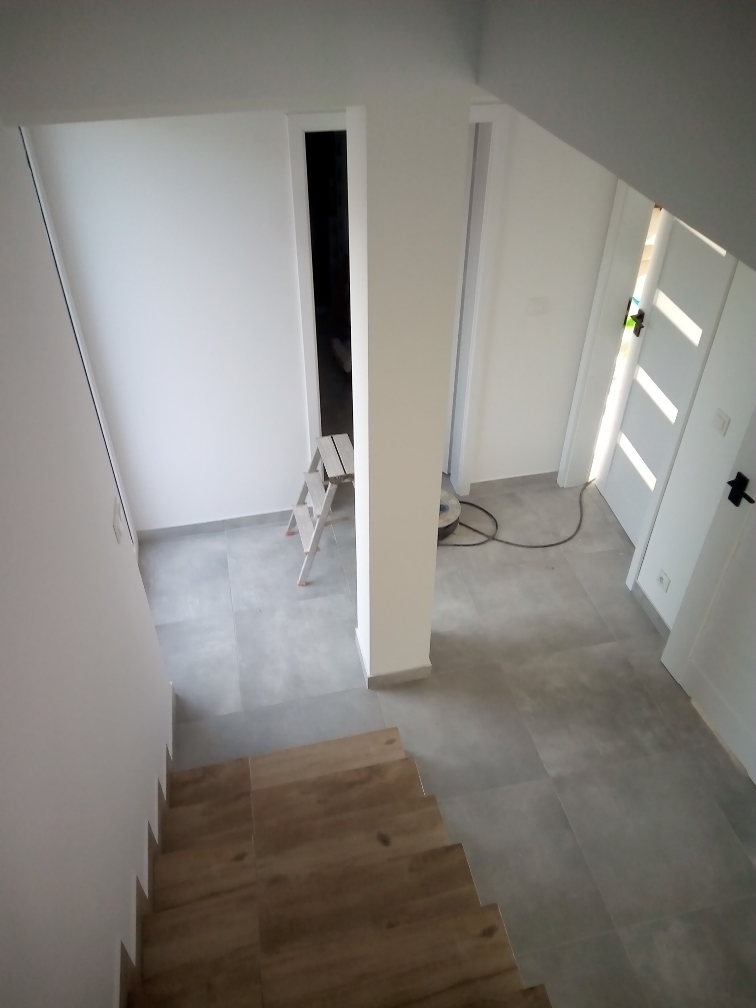 Wnętrze domu w trakcie wykańczania: szare płytki podłogowe, drewniane schody, drabina malarska i sprzęt do czyszczenia podłóg widoczne z góry.