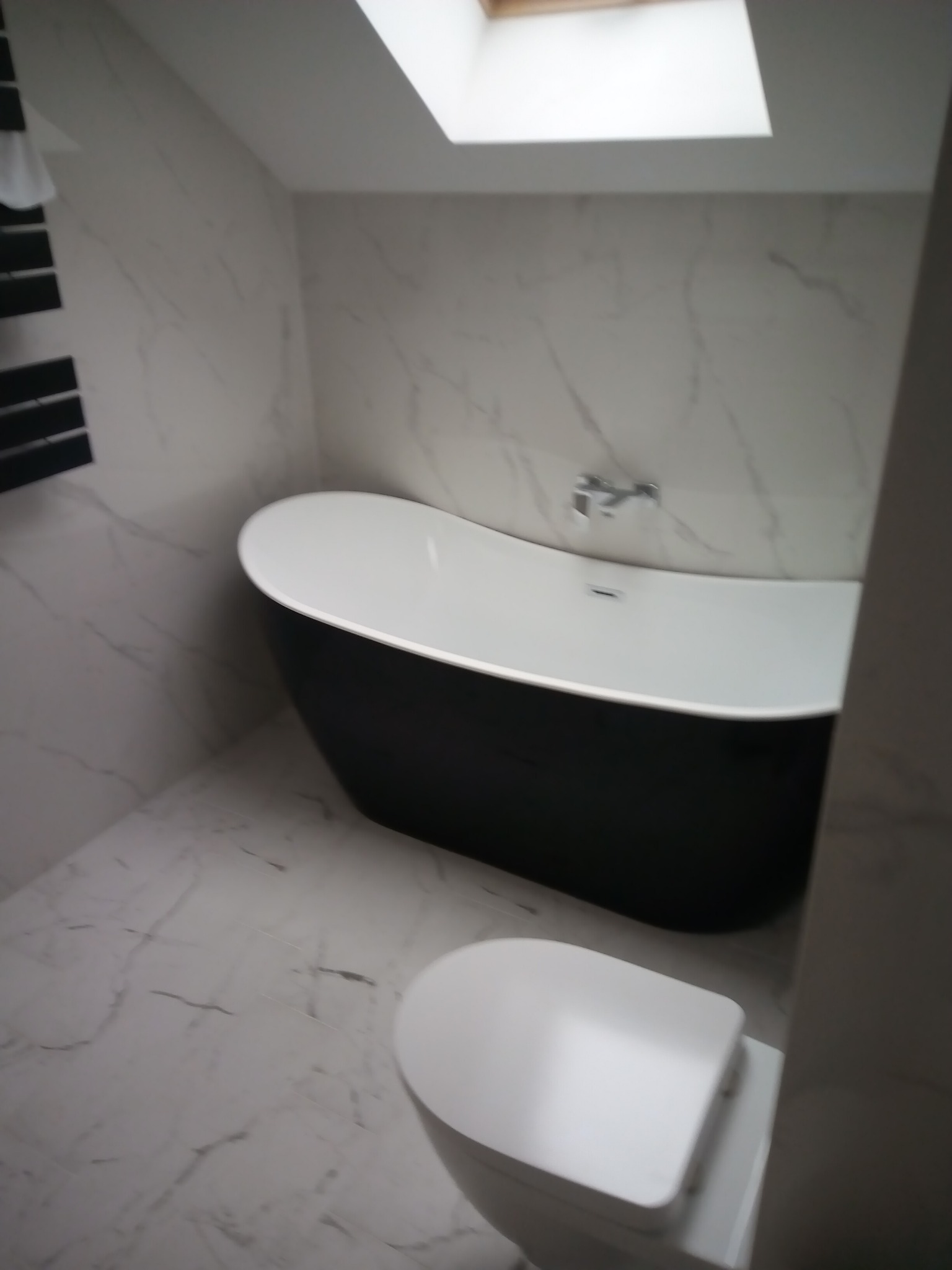 Czarno-biała wolnostojąca wanna obok toalety w łazience wykończonej białym marmurem, oświetlonej przez okno dachowe.