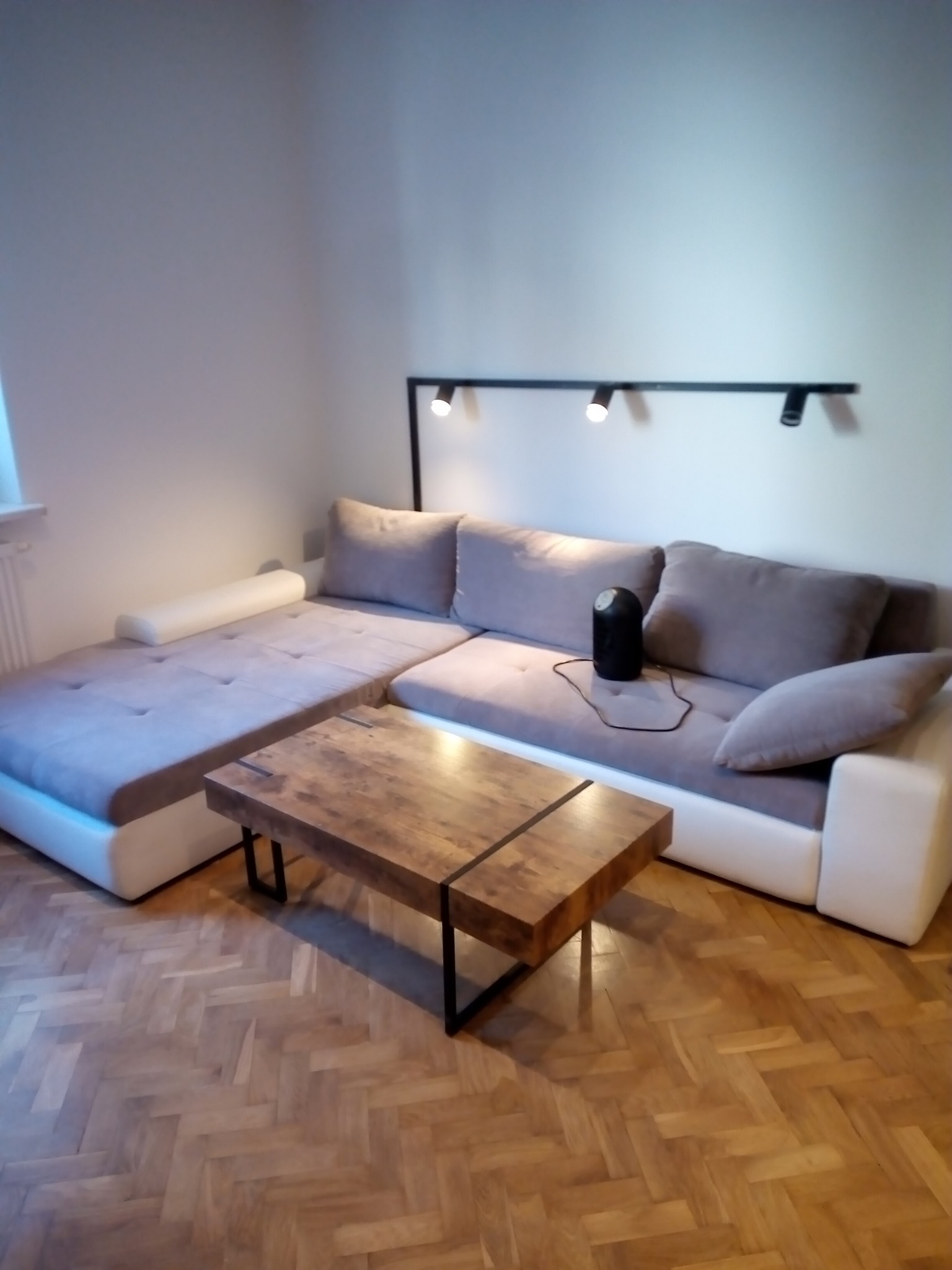 Jasnoszara narożna sofa z drewnianym stolikiem kawowym na parkiecie w jodełkę, oświetlona trzema punktowymi lampami na ścianie.