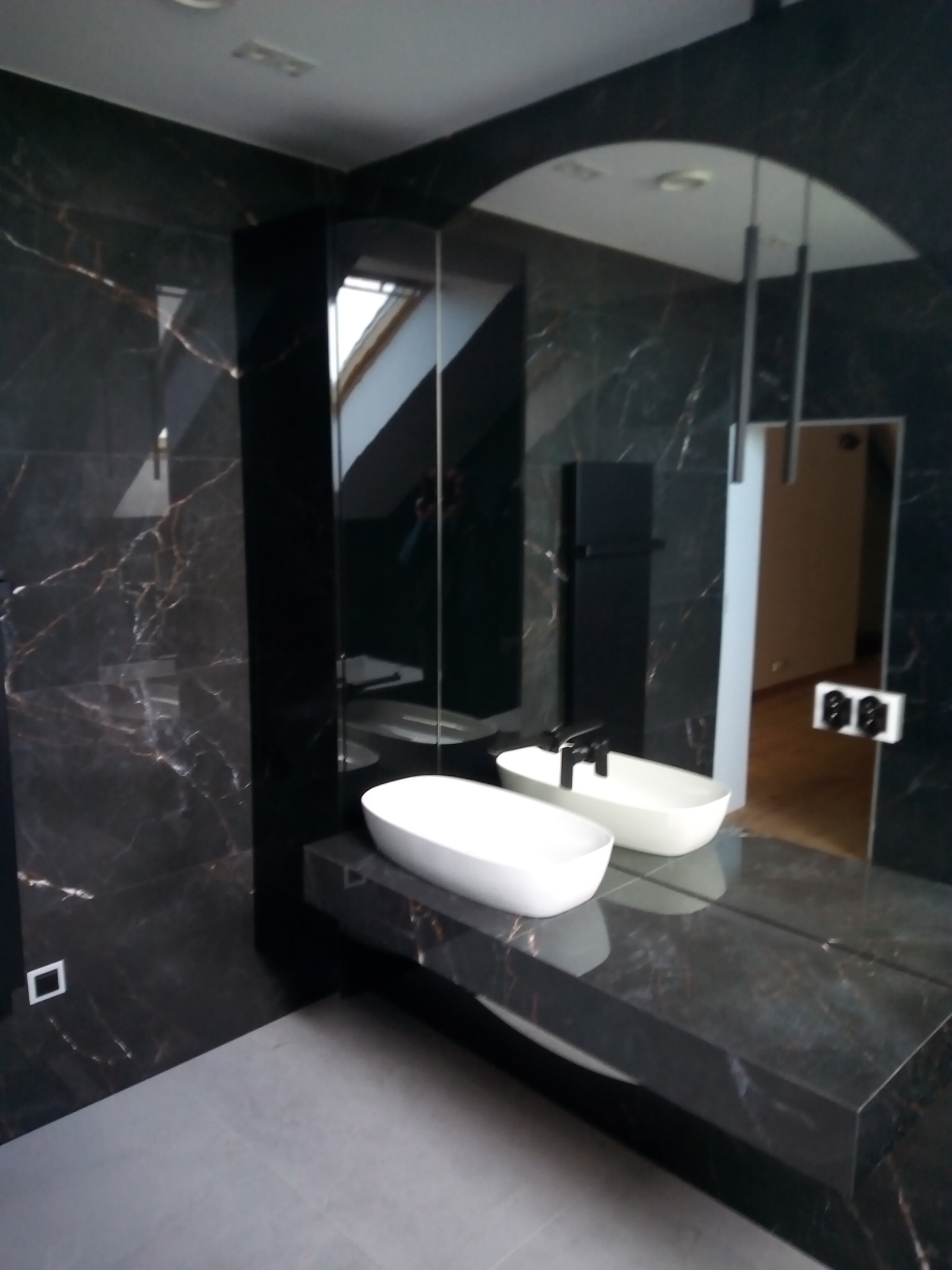 Nowoczesna łazienka z czarnymi marmurowymi płytkami, podwójną białą umywalką nablatową i lustrem z oświetleniem punktowym, widok na drzwi do innego pomieszczenia.