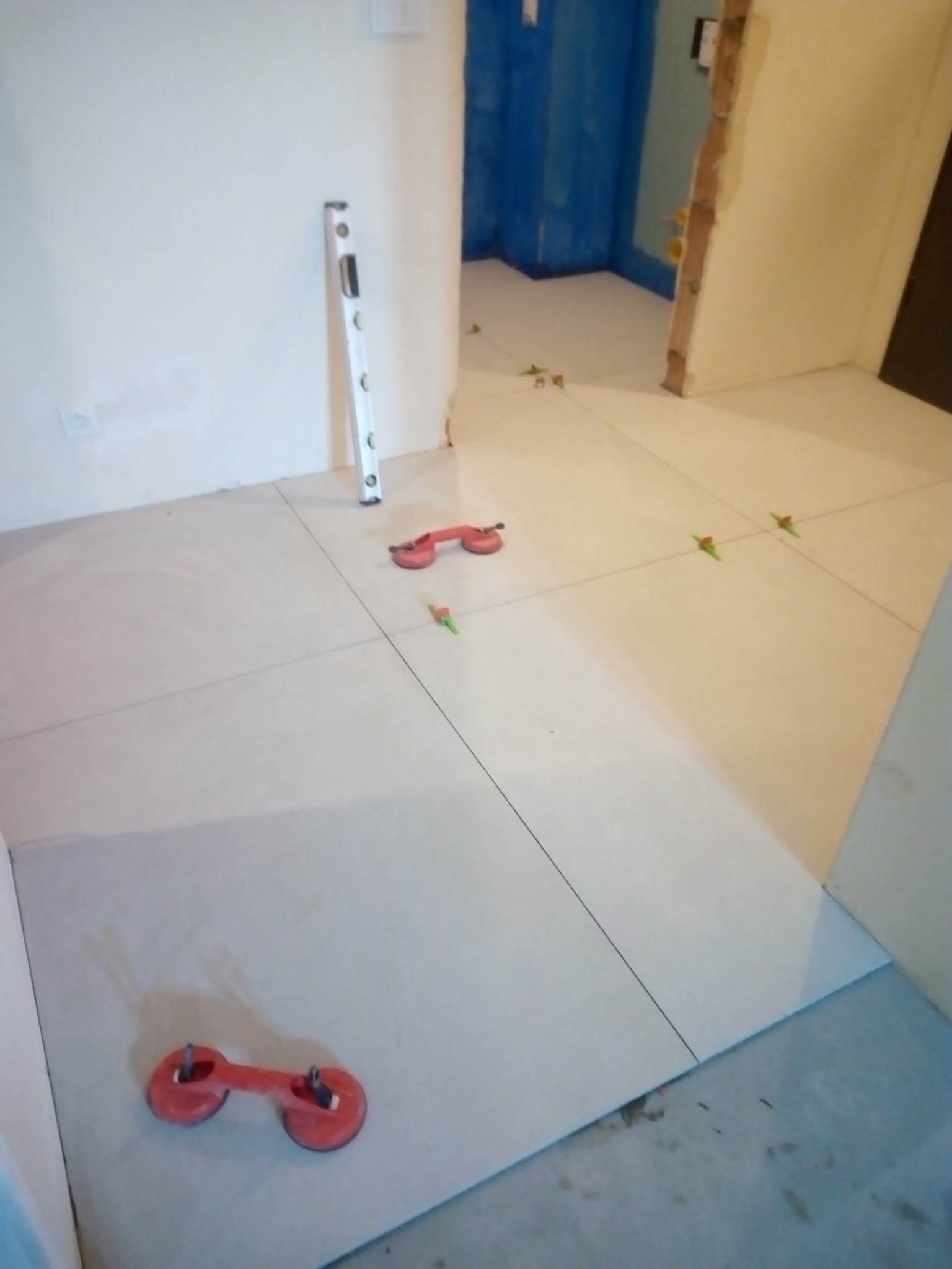 Układanie jasnych płytek podłogowych, widoczne przyssawki do przenoszenia, poziomica i krzyżyki dystansowe w trakcie prac wykończeniowych.