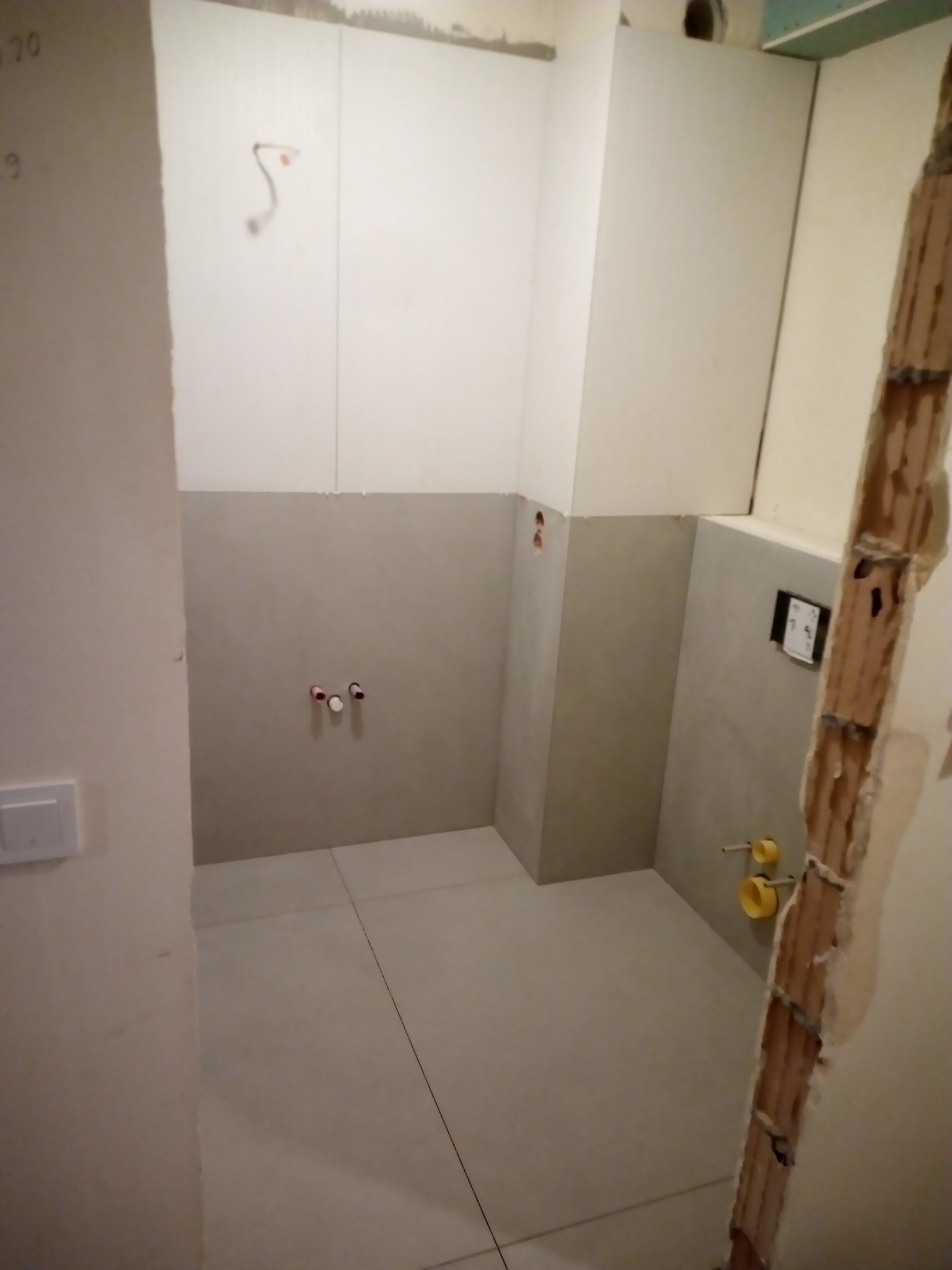 Wnętrze łazienki w trakcie prac wykończeniowych, widoczne położone płytki podłogowe, częściowo ułożone płytki ścienne, wystające rury i instalacje, oraz odsłonięta cegła w rogu pomieszczenia.