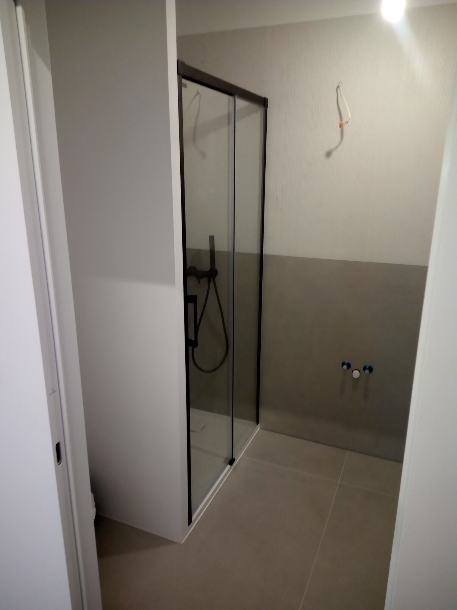 Wnętrze łazienki w trakcie remontu, widoczna kabina prysznicowa z czarnymi profilami, ściana z częściowo położonymi szarymi płytkami, wystające kable i rury, surowy stan wykończenia.
