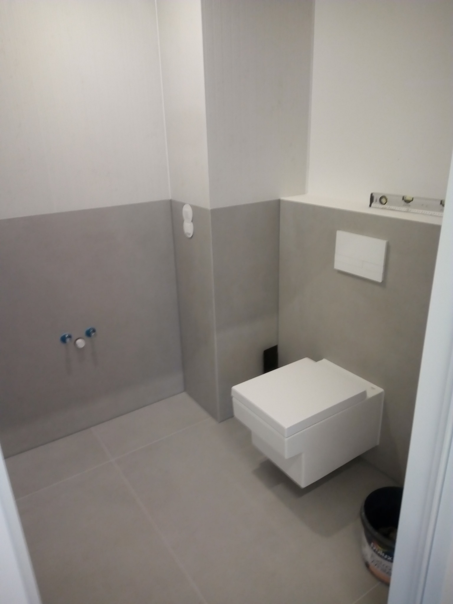 Podwieszana toaleta z zabudowaną spłuczką w szarej łazience, widoczny poziomica na ścianie i wiadro z farbą Dulux