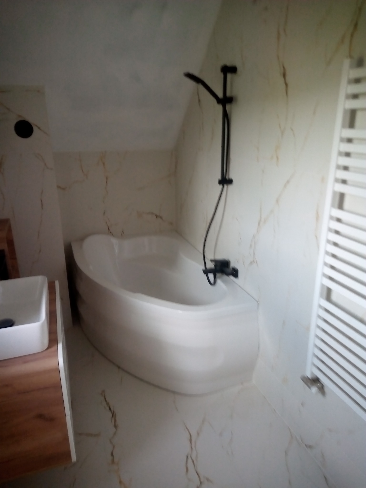 Nowoczesna łazienka na poddaszu z białą wanną asymetryczną, czarną armaturą i grzejnikiem drabinkowym na ścianie wyłożonej płytkami imitującymi marmur.