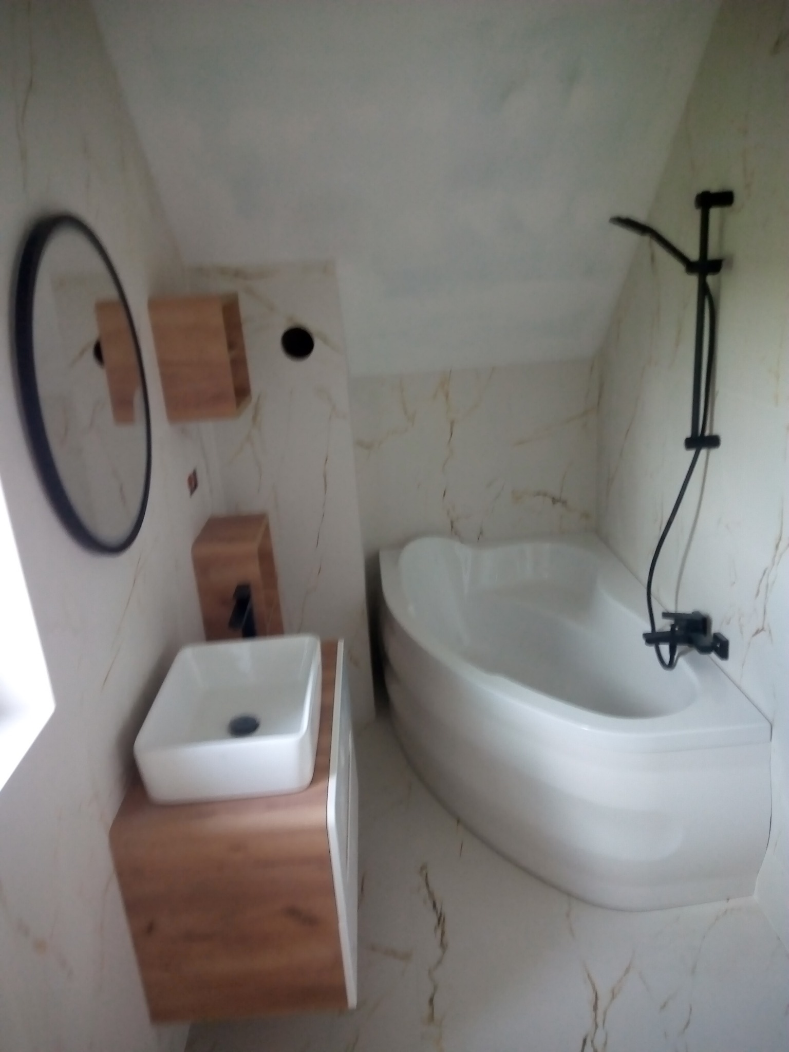 Ukośna łazienka z wanną narożną, białą umywalką na drewnianej szafce, czarną armaturą i marmurkowymi płytkami.