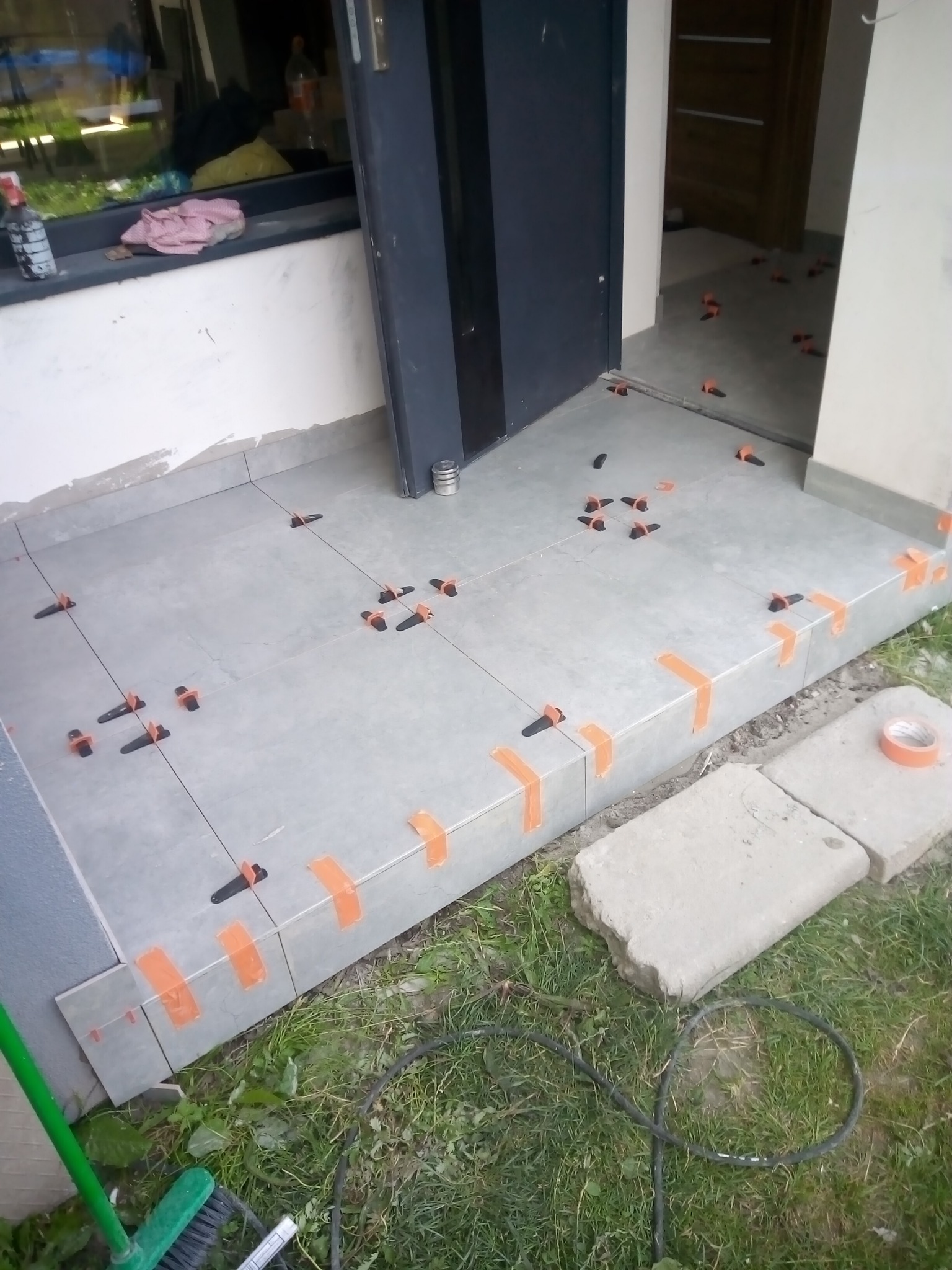 Układanie szarych płytek na schodach wejściowych z użyciem pomarańczowej taśmy i czarnych klipsów poziomujących, widoczne narzędzia i fragment trawnika.