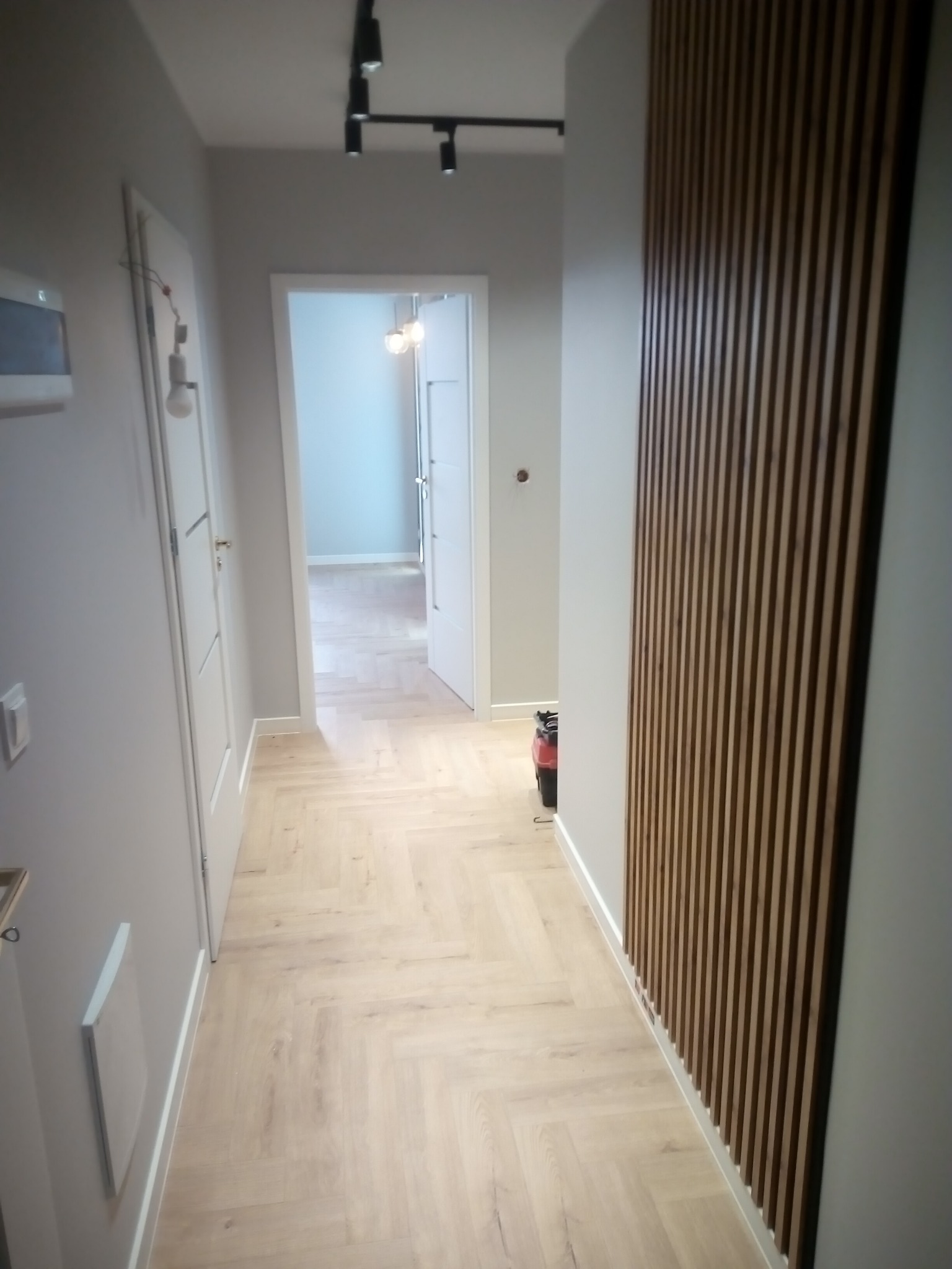 Nowoczesny korytarz z drewnianą okładziną ścienną z pionowych listew, jasną podłogą ułożoną w jodełkę, białymi drzwiami i oświetleniem szynowym na suficie.