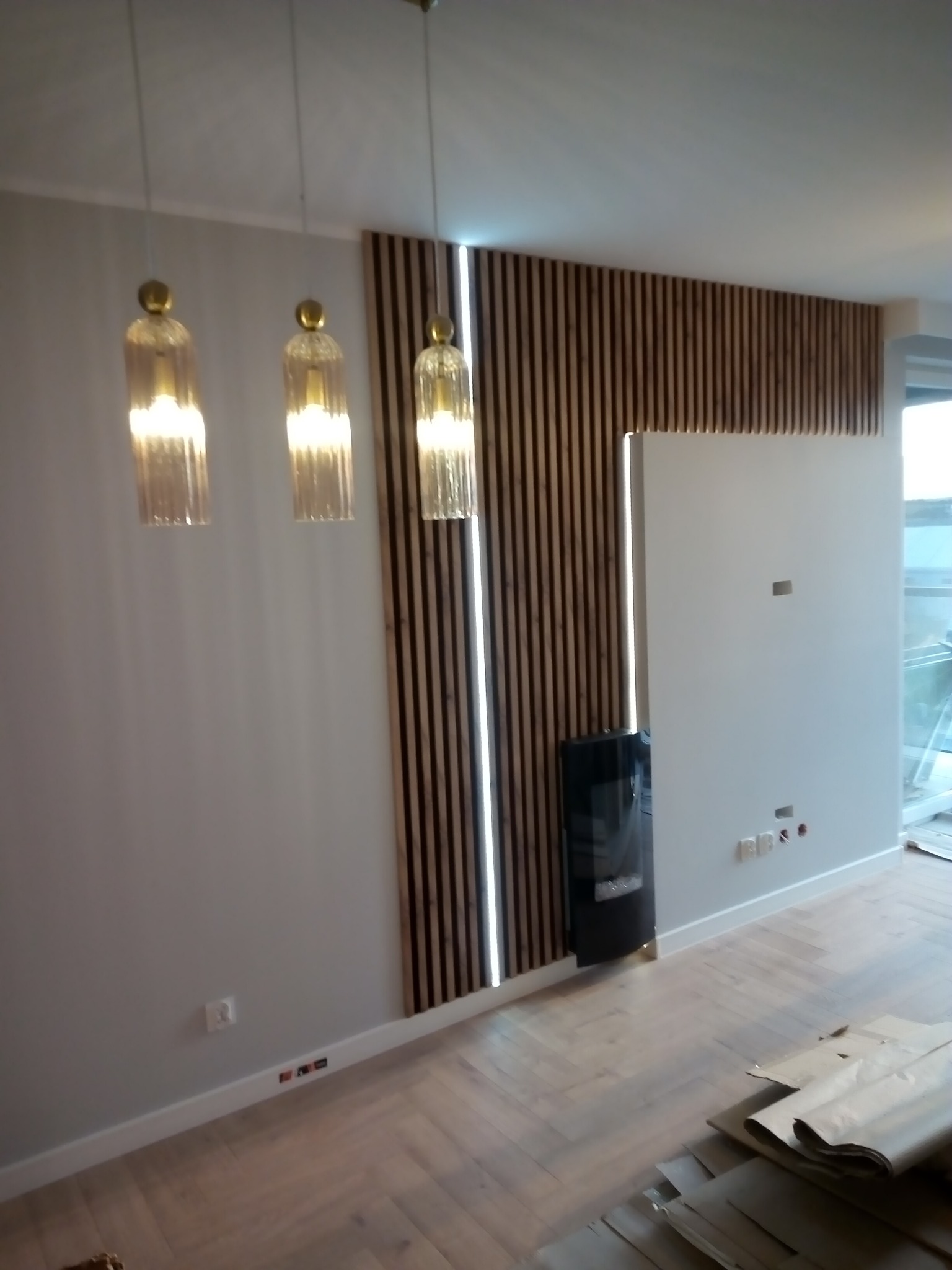 Ściana z drewnianymi lamelami, oświetleniem LED i kominkiem elektrycznym. Na pierwszym planie podłoga z parkietem w jodełkę oraz materiały budowlane.