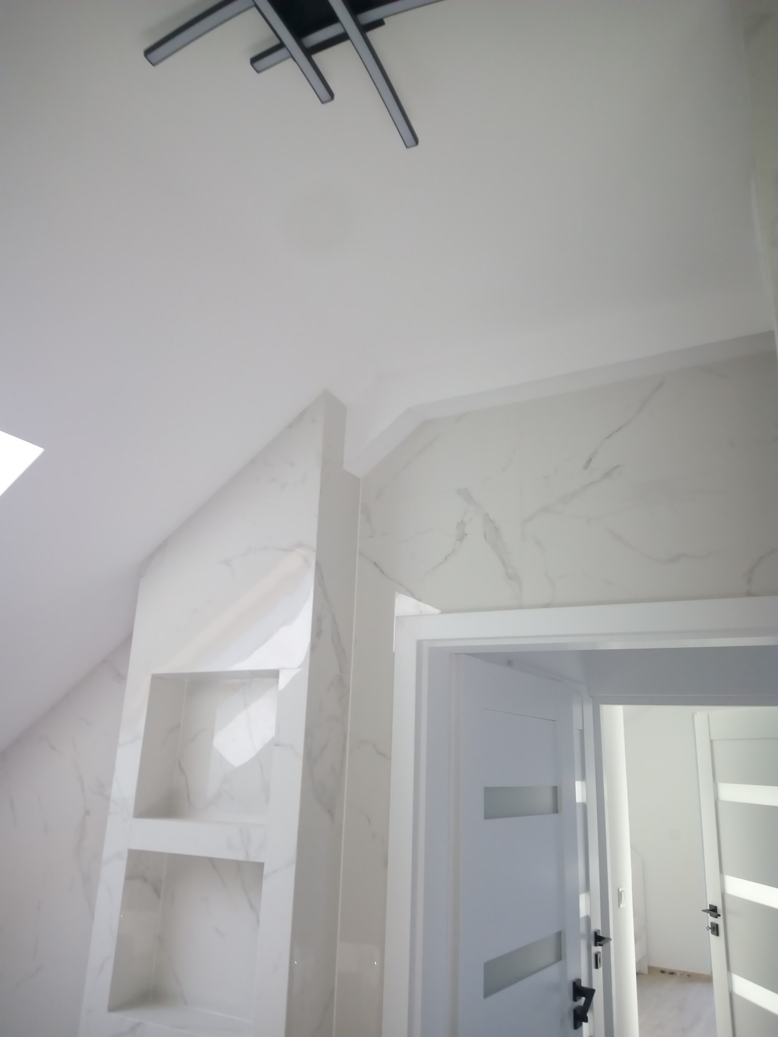 Wykończona łazienka w bieli z marmurowymi płytkami, wbudowane półki w ścianie, nowoczesna lampa sufitowa i widok na drugie pomieszczenie przez otwarte drzwi.