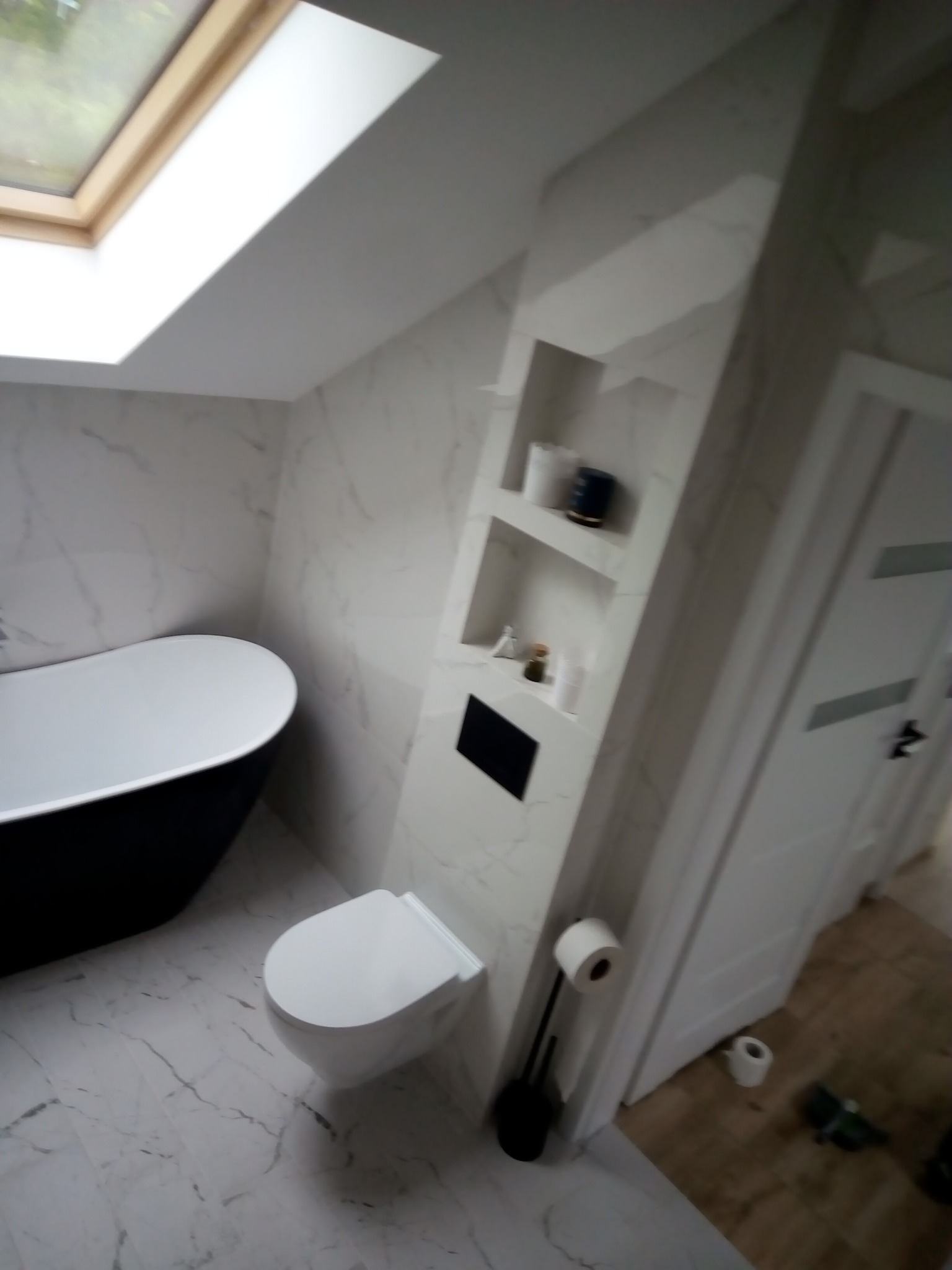Nowoczesna łazienka na poddaszu z biało-szarą marmurową okładziną ścian i podłogi, czarną wolnostojącą wanną, podwieszaną toaletą i wnęką z półkami.