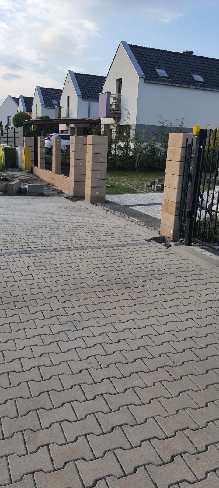 Wznoszenie ogrodzenia z pustaków betonowych, widoczne słupki i fragment muru, kostka brukowa na podjeździe, w tle domy jednorodzinne
