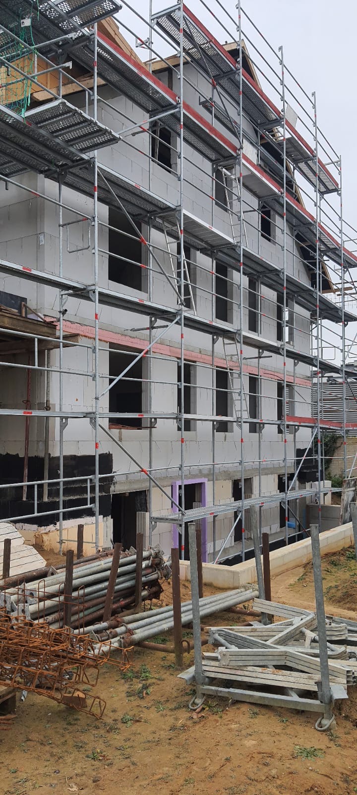Wielopiętrowy budynek w trakcie wznoszenia ścian z bloczków, otoczony rusztowaniem, z widocznymi elementami konstrukcyjnymi i materiałami budowlanymi na ziemi.