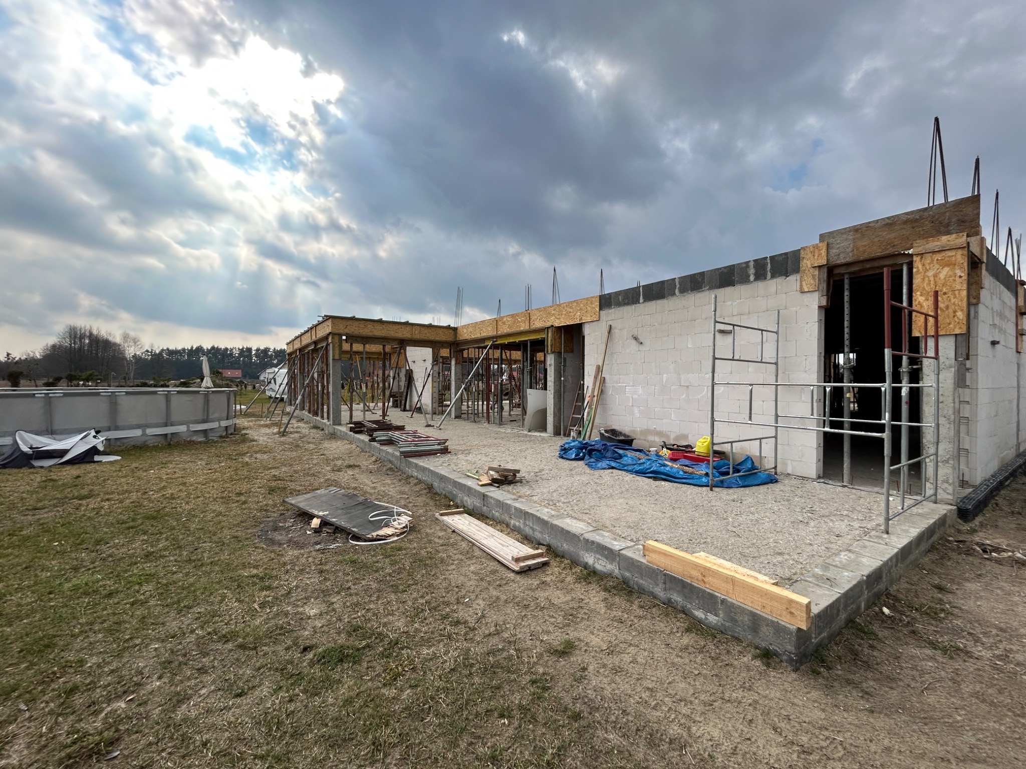 Budowa domu jednorodzinnego w trakcie murowania ścian z bloczków betonowych, widoczne zbrojenia, szalunki i przygotowany teren wokół budynku.