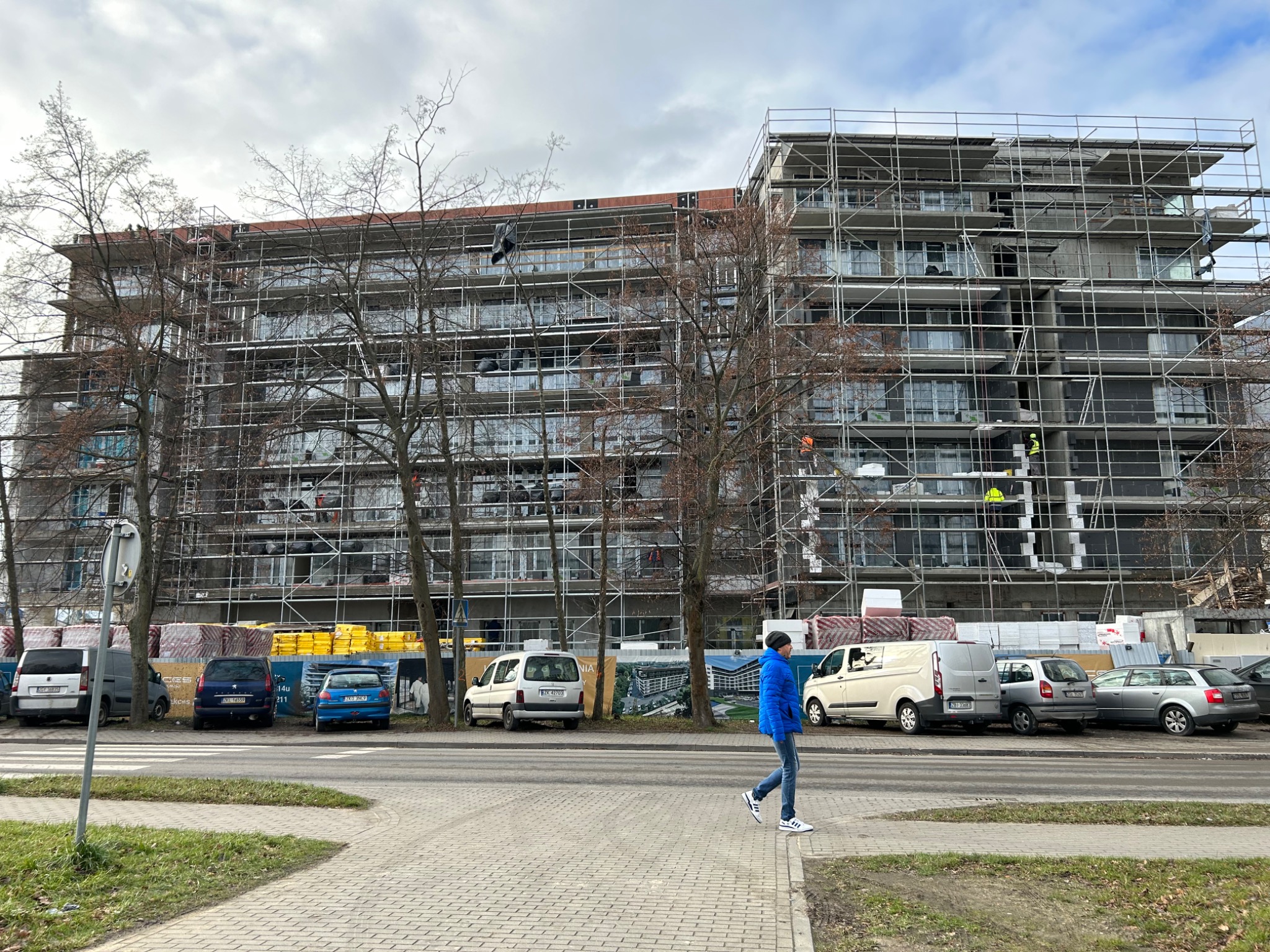 Wielopiętrowy budynek w trakcie prac budowlanych, otoczony rusztowaniami, z widocznymi pracownikami w oddali i zaparkowanymi samochodami wzdłuż ulicy, na pierwszym planie spacerujący przechodzień.
