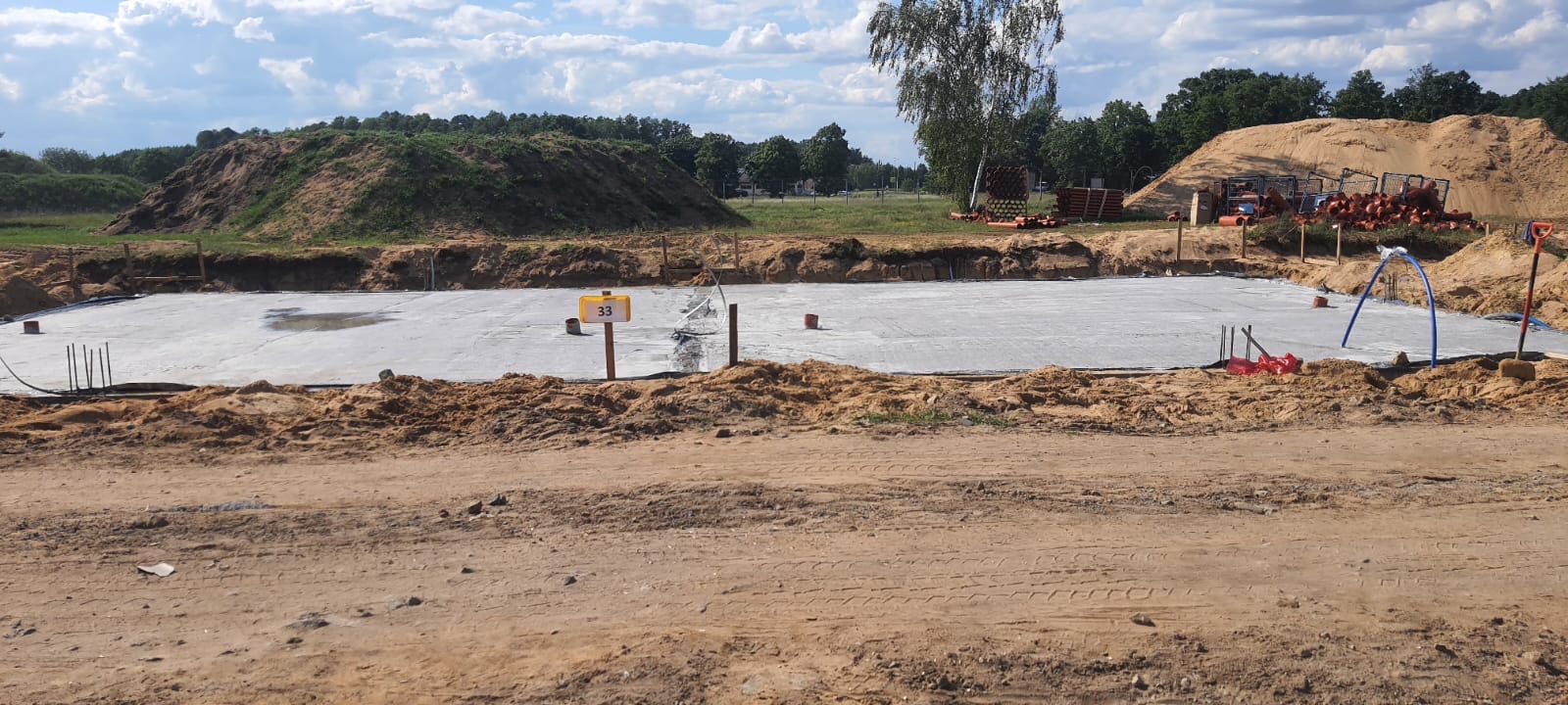 Wylany fundament pod budynek z widocznymi wystającymi prętami zbrojeniowymi, numerem działki 33 i stertami ziemi w tle.